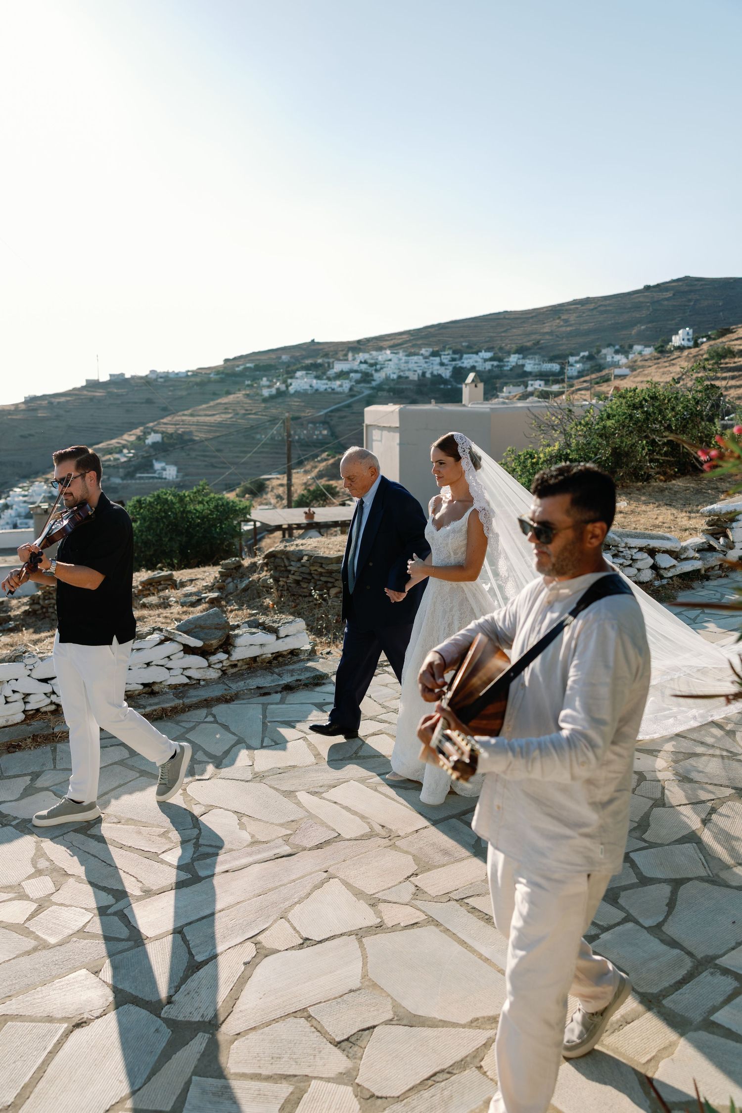 Bride in white veil walks with guests as musicians play guitar and violin at an outdoor Greek island wedding ceremony.