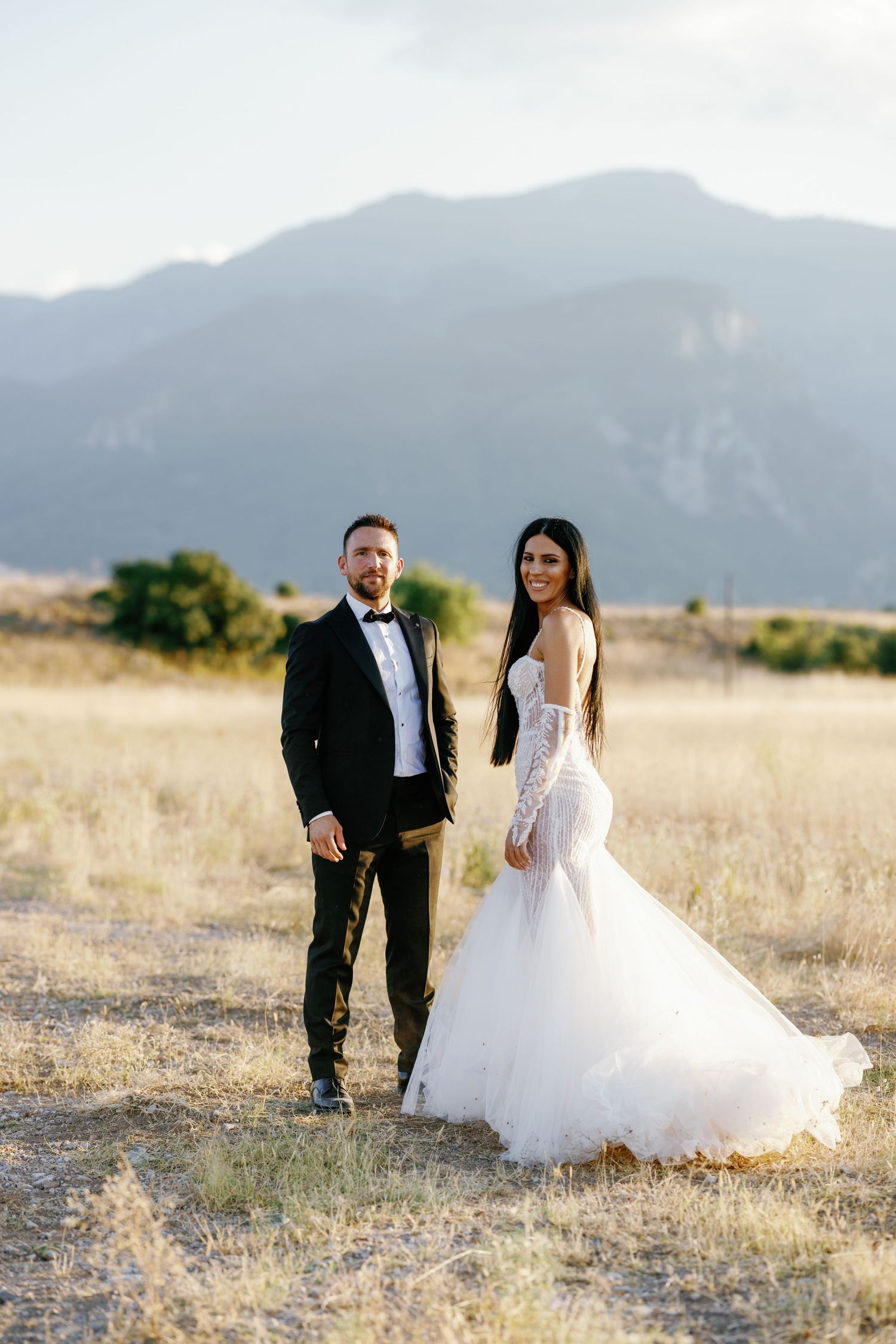 Newlyweds pose together in field with mountain backdrop at sunset.