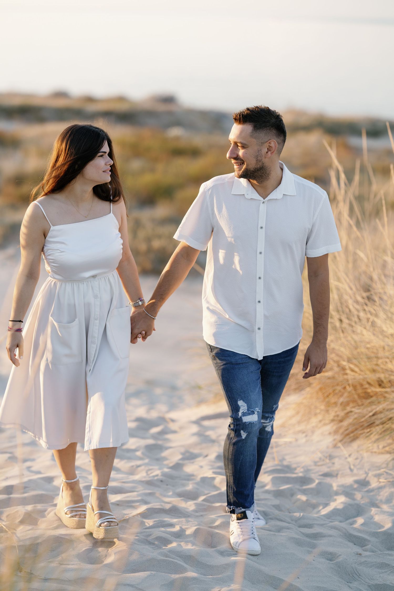 Couple walking hand in hand on sandy beach wearing coordinated white outfits.