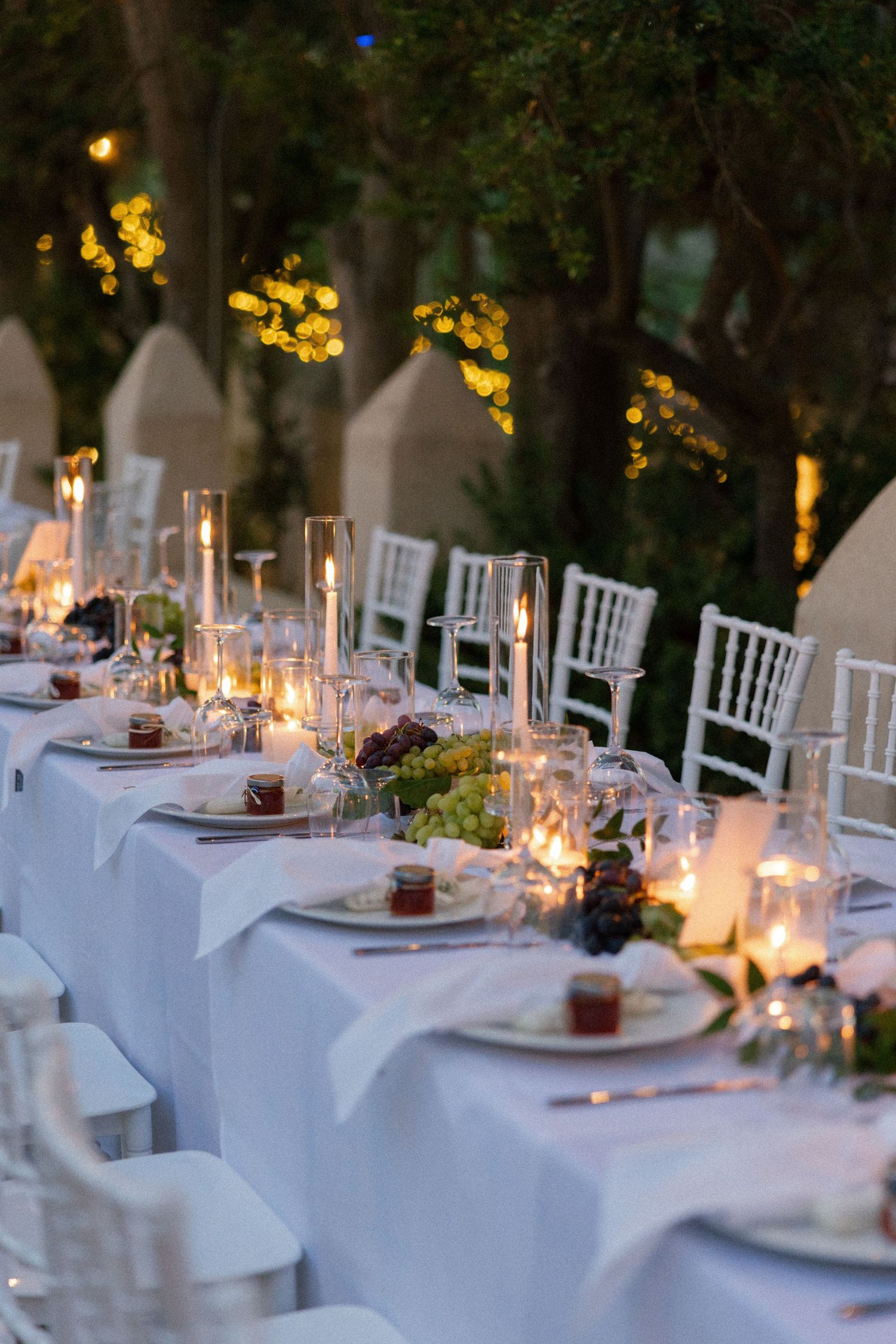 Elegant outdoor wedding reception with long white-draped tables, candles, and white chiavari chairs set under trees at dusk.