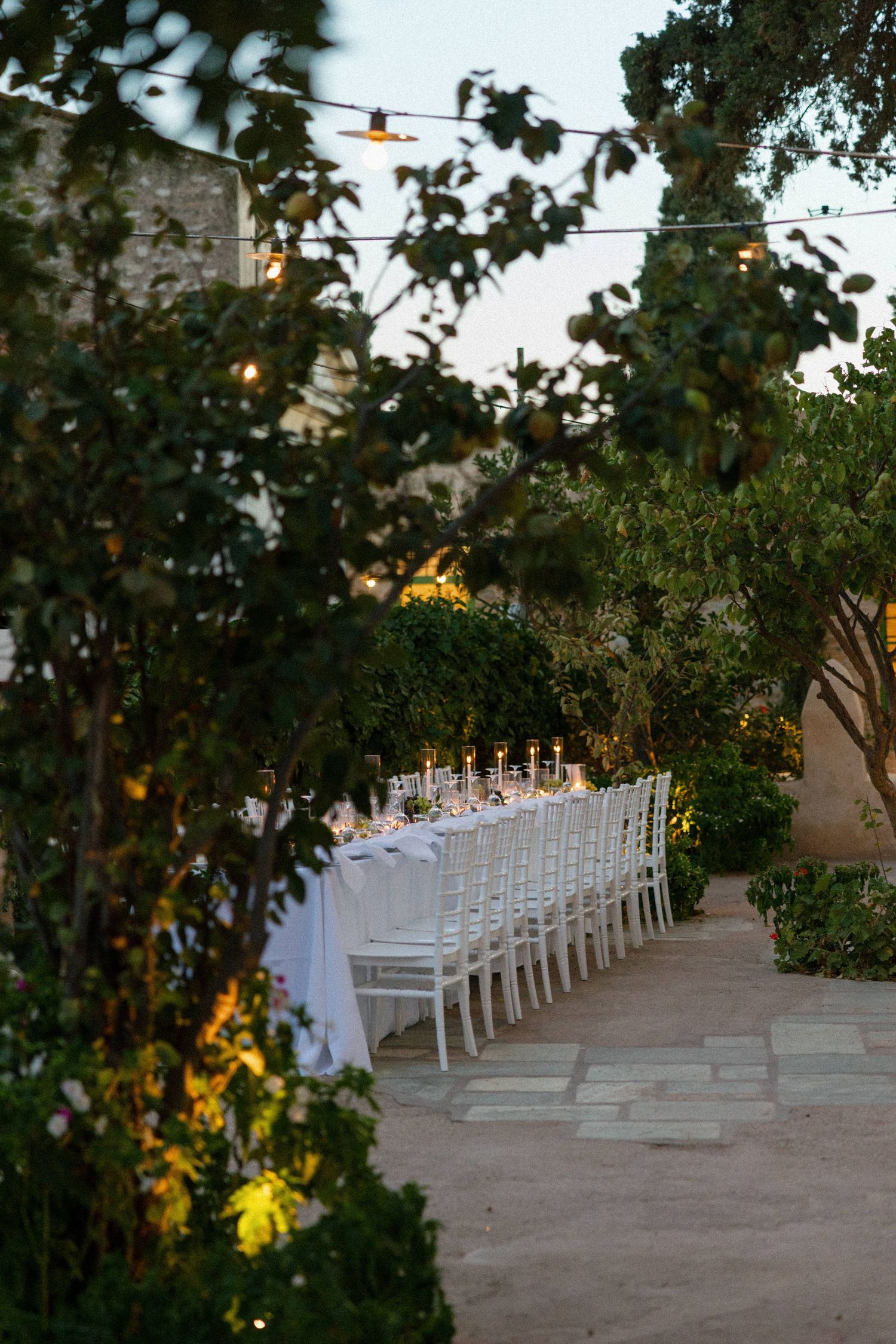 Elegant outdoor wedding reception with long white table, candles, white chairs, string lights, and lush garden greenery at dusk.
