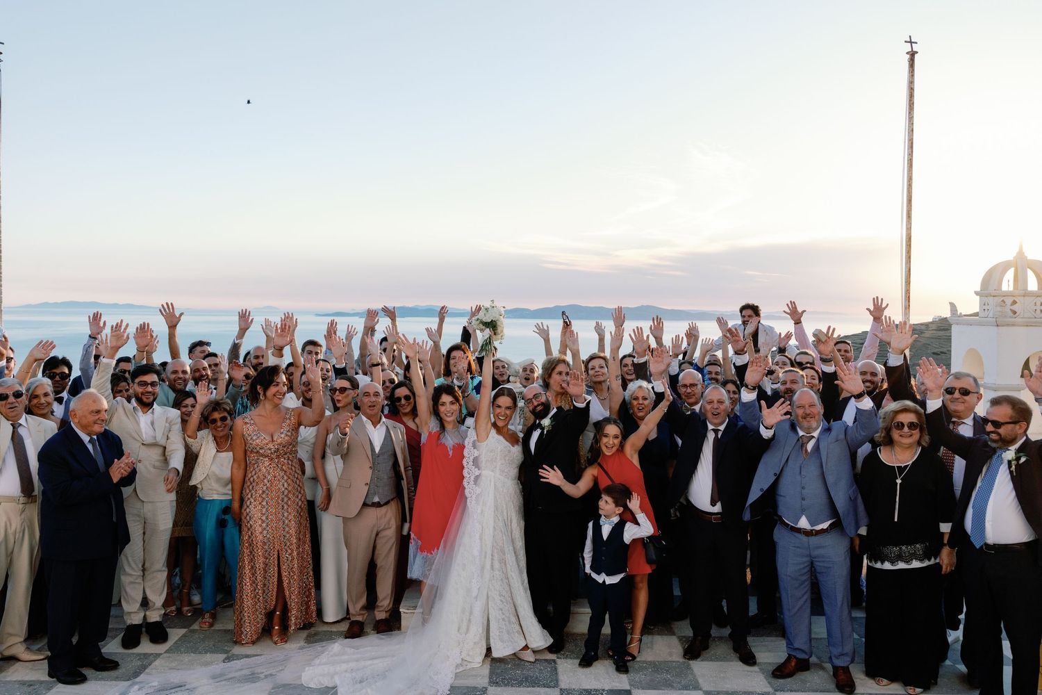 Large wedding group photo with bride and groom, guests cheering with raised arms at an outdoor seaside venue at sunset.