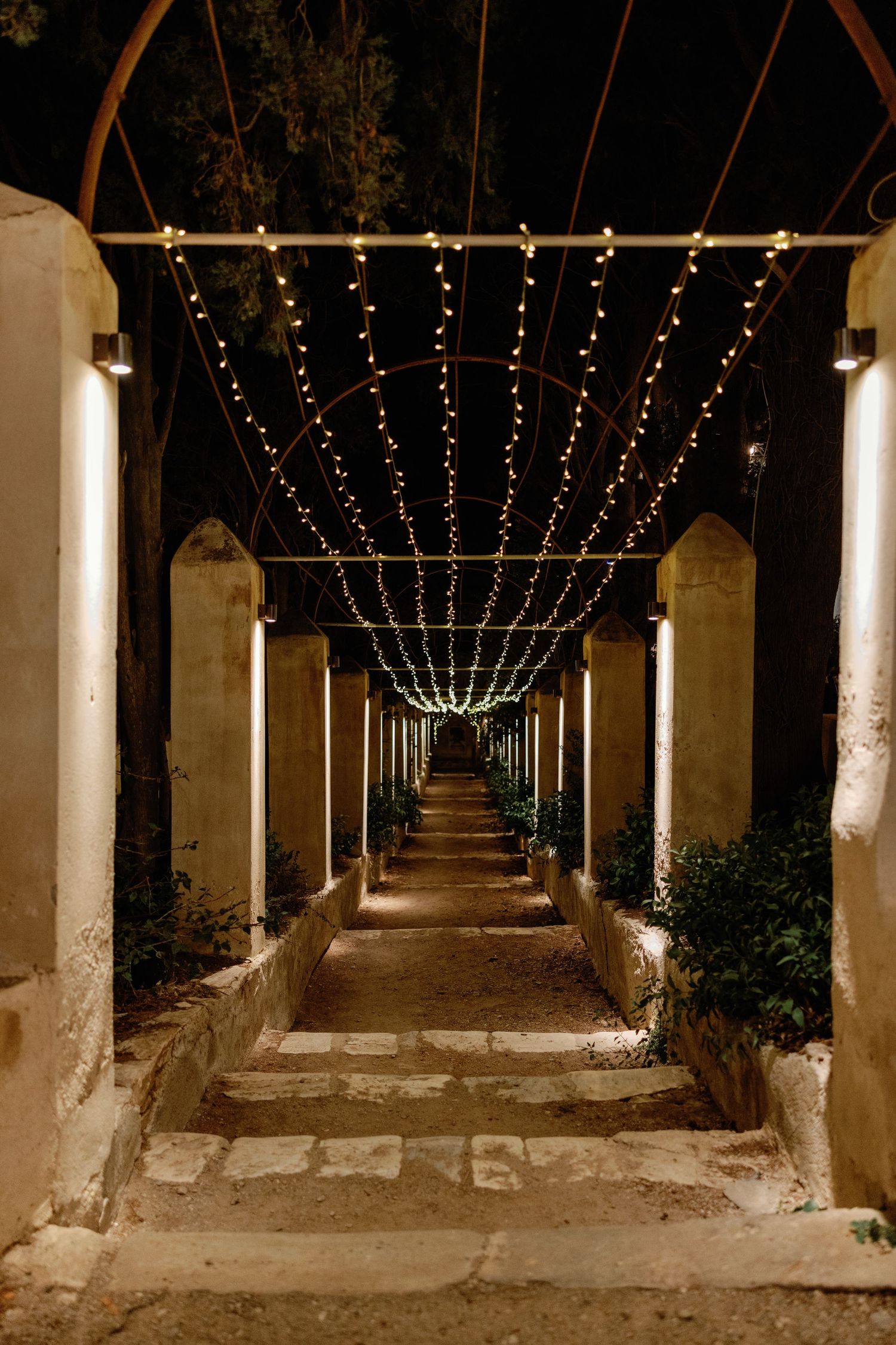 A romantic outdoor corridor with string lights overhead, stone columns, and steps leading into the night.