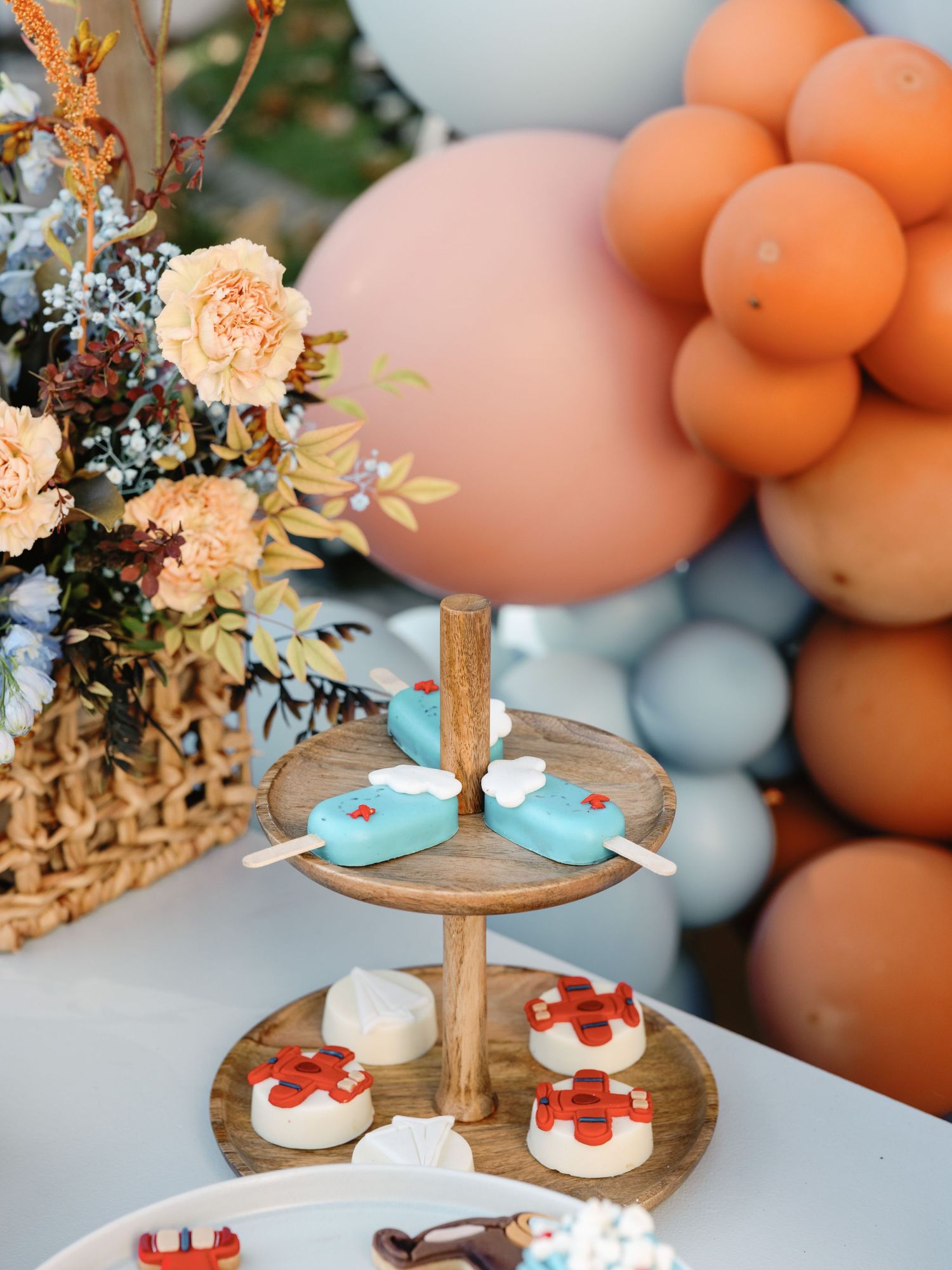 Elegant dessert display with wooden tiered stands adorned with treats against a peach and blue balloon backdrop.