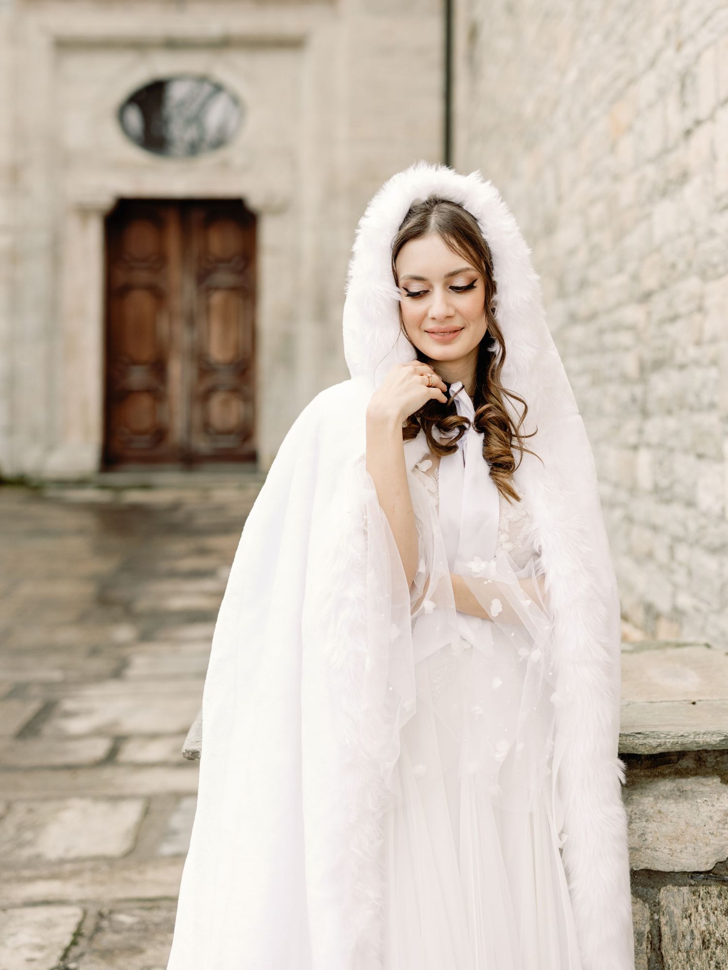 A bridal portrait featuring an elegant white wedding gown and flowing veil against a stone wall backdrop.