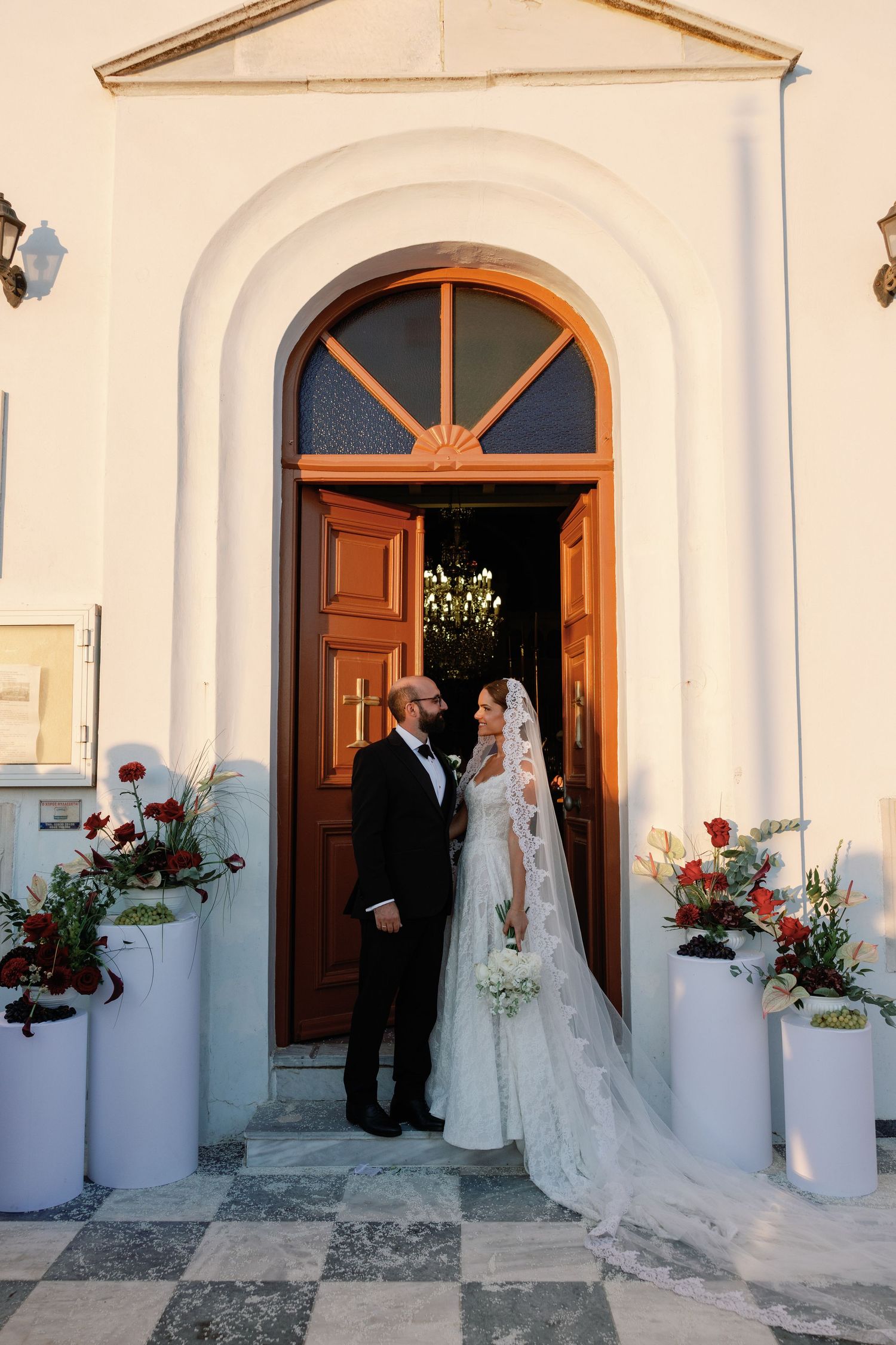 Bride and groom stand together at ornate church entrance with floral arrangements and checkered floor on their wedding day.