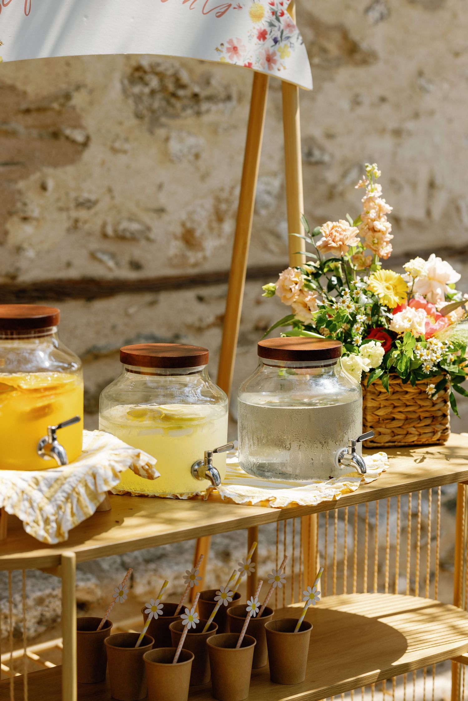 Beverage station with glass dispensers and floral arrangements on wooden shelving.