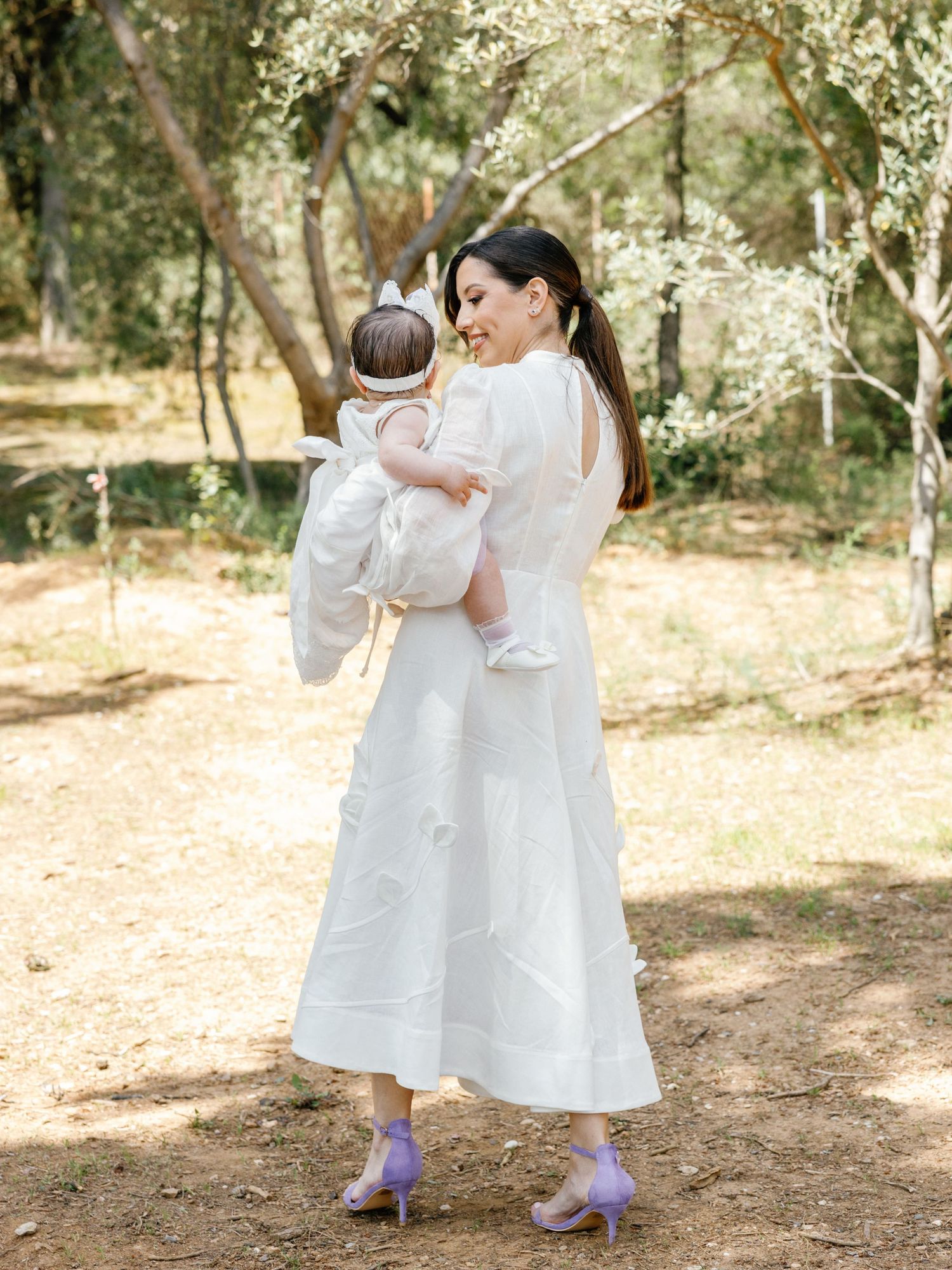 A woman in a flowing white dress and purple shoes holds a baby while standing in a sunlit wooded area.