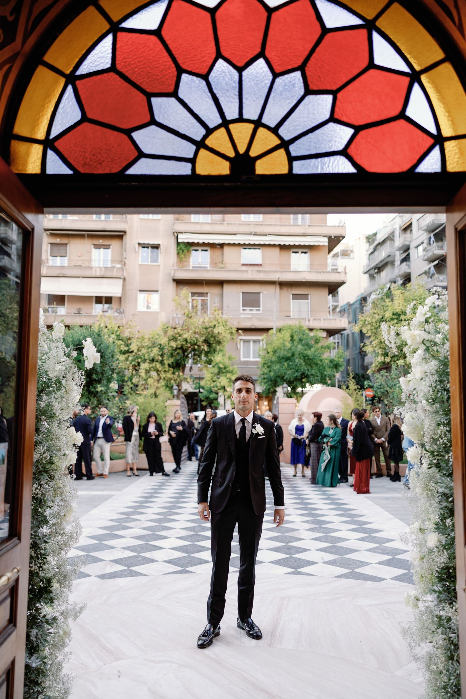 Wedding guests gather in an elegant courtyard with a checkered floor pattern and decorative archway.