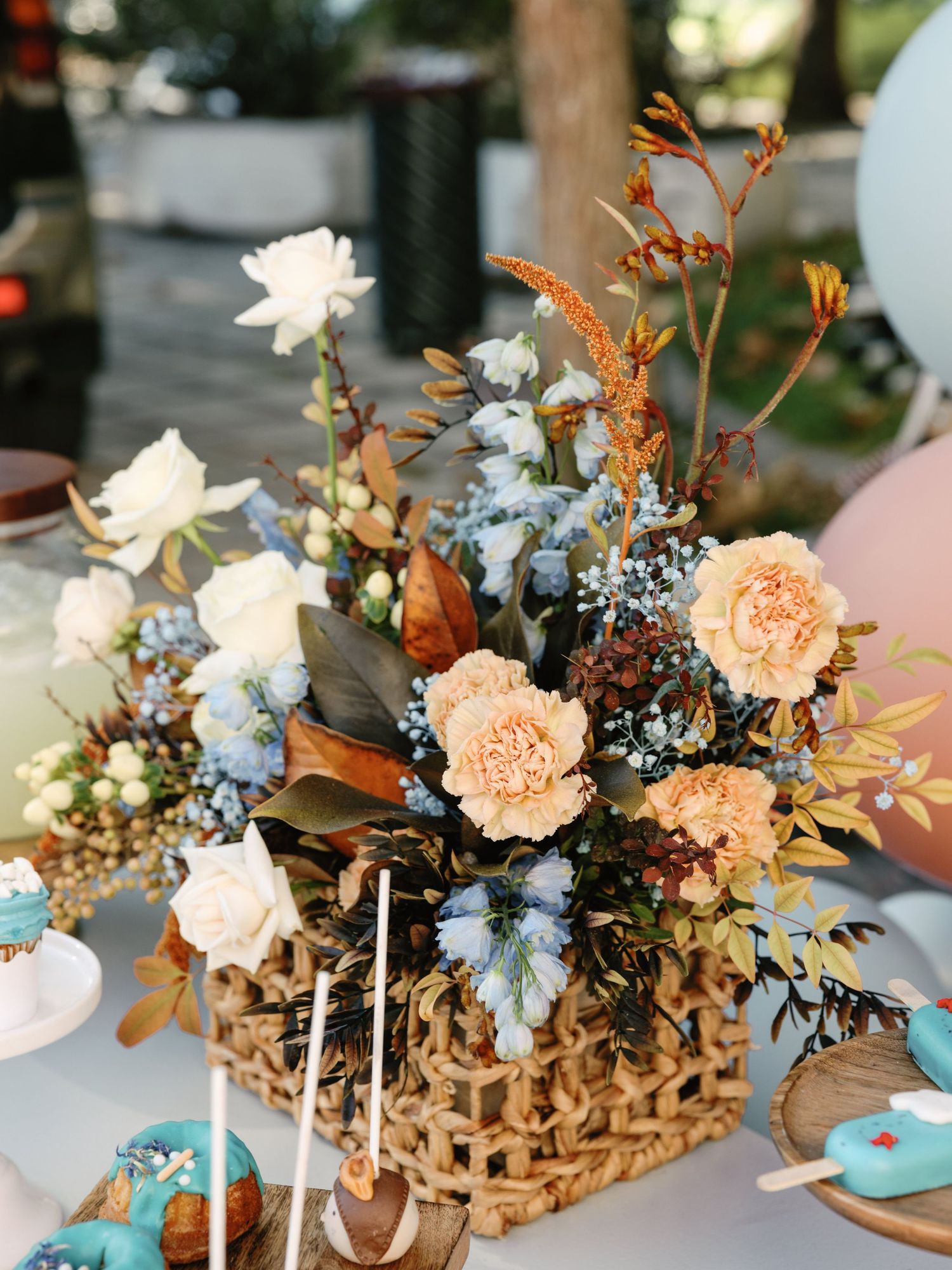 Rustic woven basket centerpiece with peach carnations, blue delphinium, and dried foliage on wooden table.