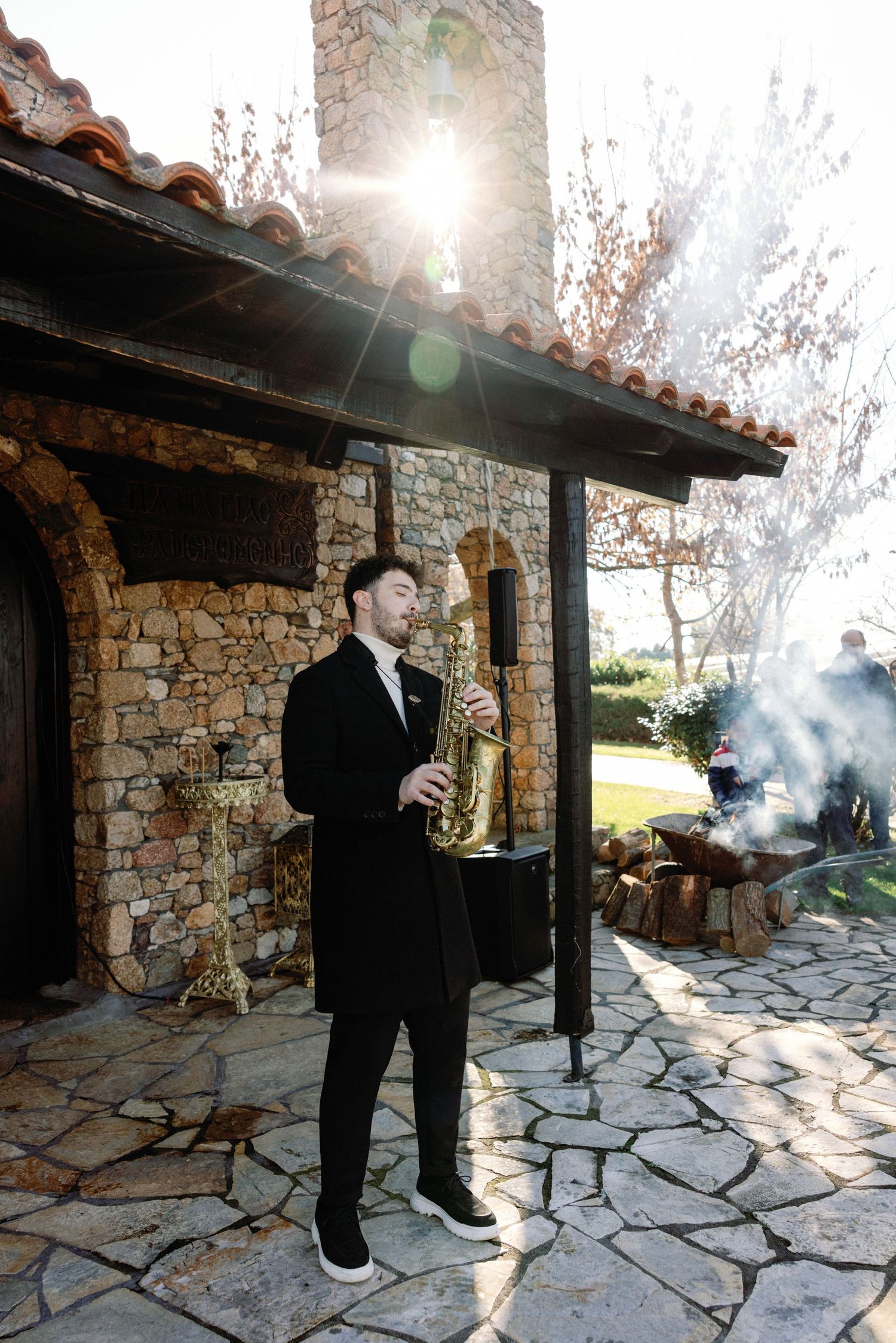 A saxophone player in formal attire performs on a stone patio with sunlight streaming through the trees.