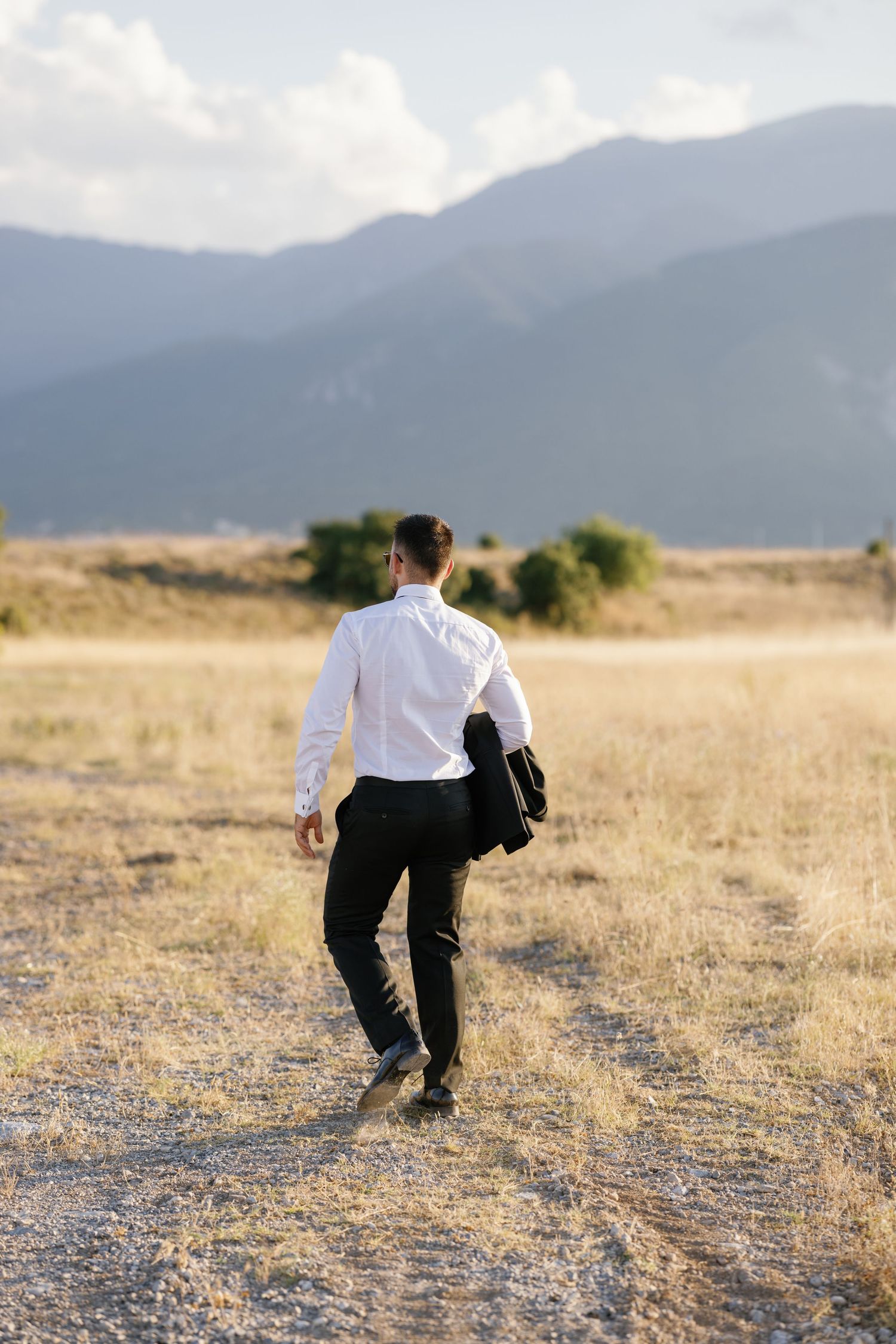 Person in formal attire walking through a golden field with mountains in the background.