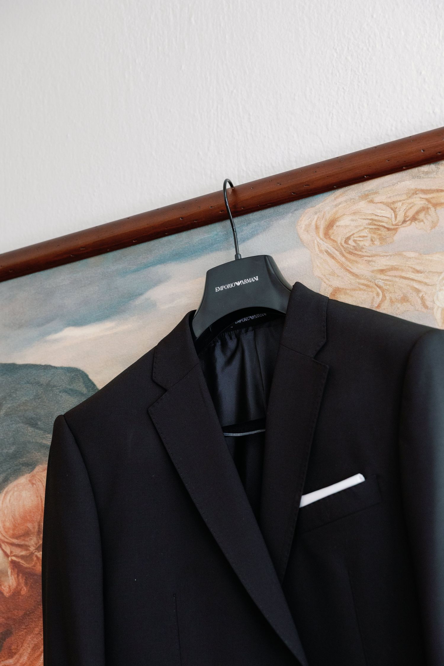 A black formal suit jacket hanging on a wooden hanger against a white wall.
