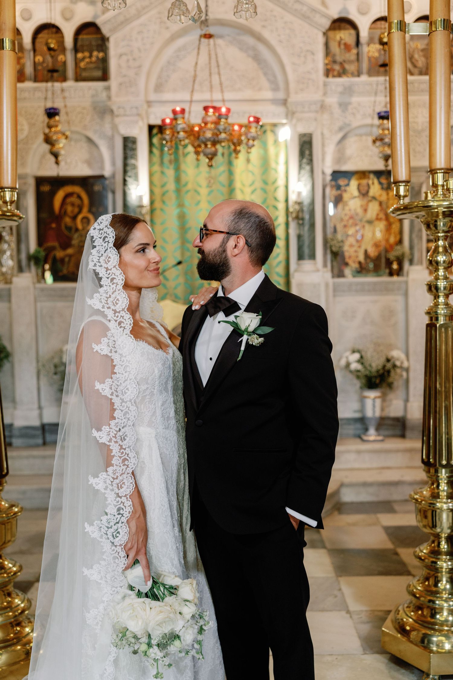 Bride and groom share a romantic moment in an ornate Orthodox church, bride wearing lace veil and groom in black tuxedo.