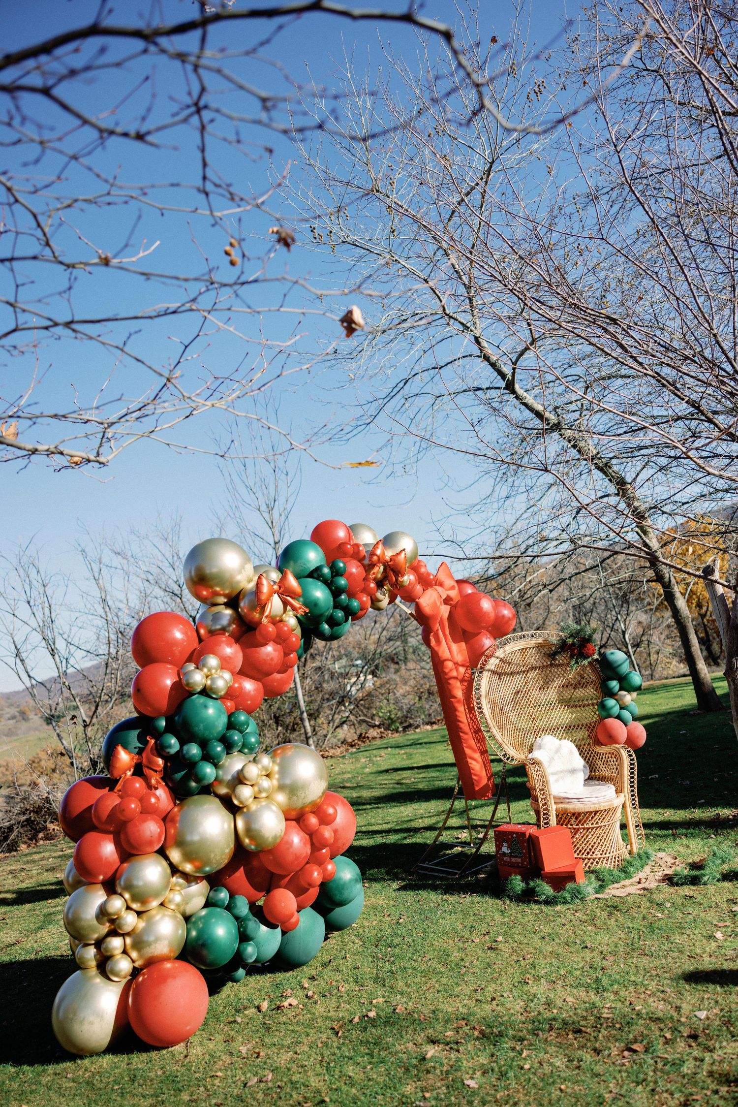 Festive outdoor balloon arch installation in progress with red, green and gold ornament-style balloons on a grassy lawn.
