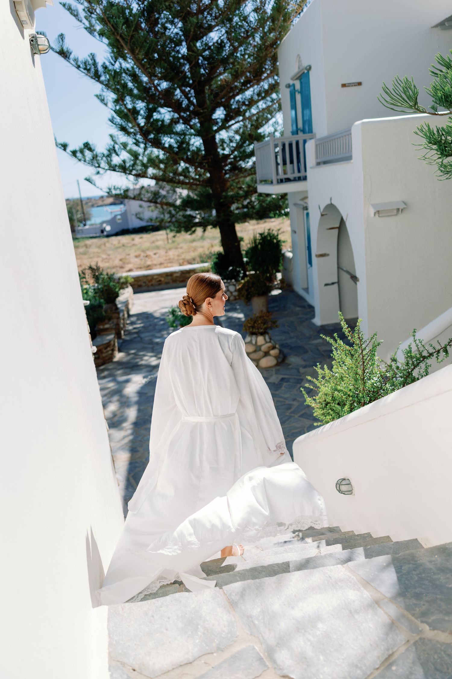Bride in flowing white gown walks up stone steps at a Greek island villa surrounded by whitewashed walls and greenery.