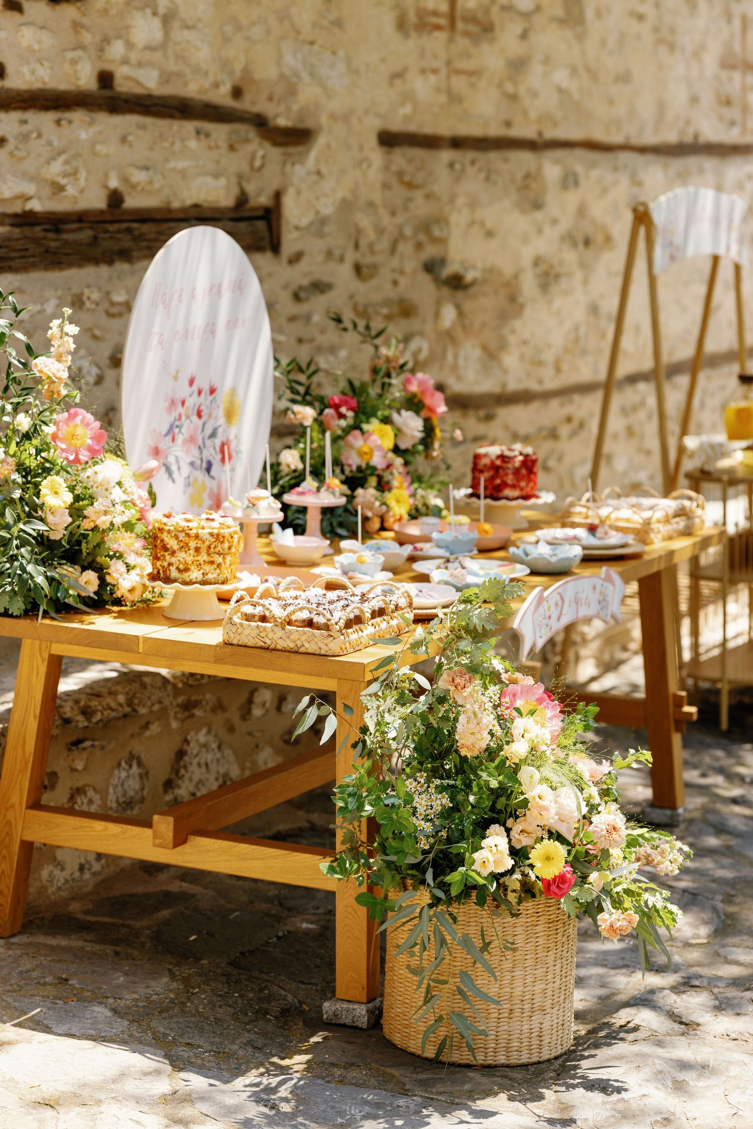 Rustic outdoor dessert table with wooden furniture and floral arrangements against a stone wall.