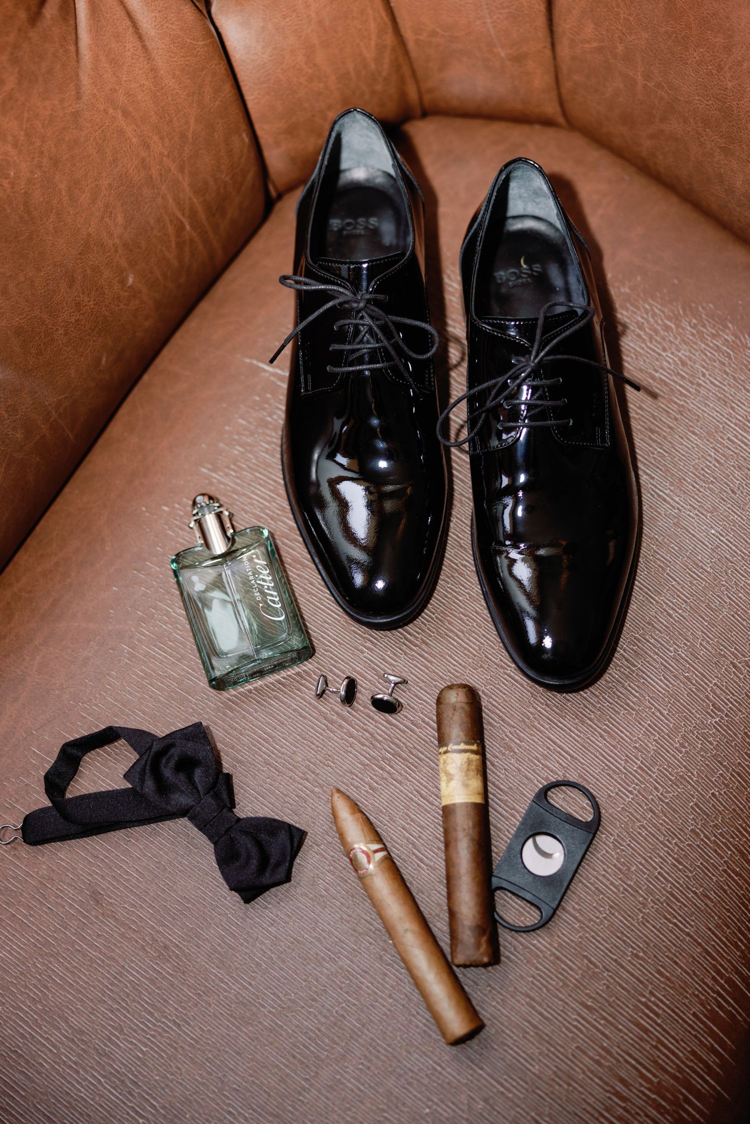Wedding accessories including black dress shoes, cologne, and bow tie arranged on brown leather.