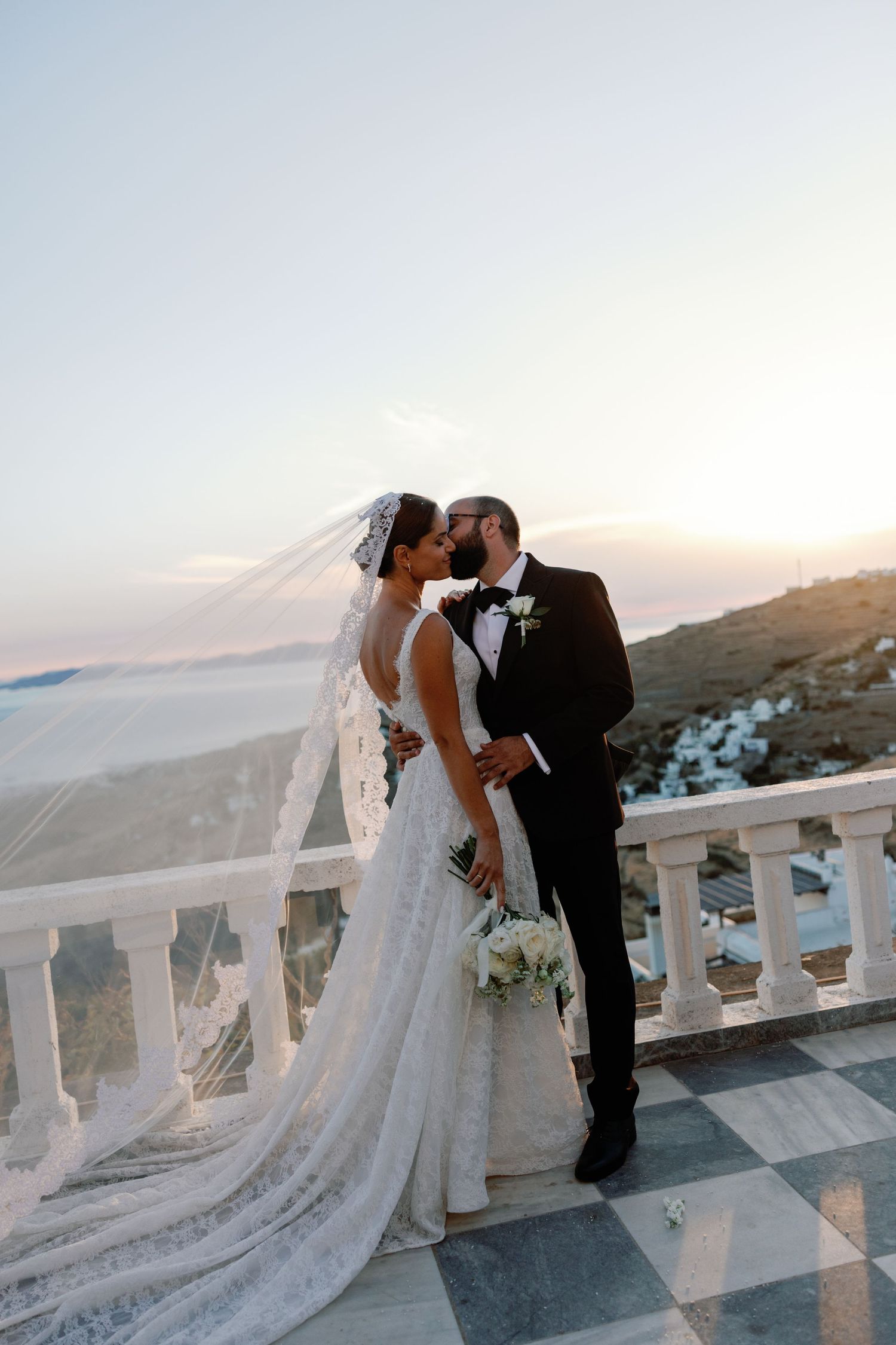 Bride and groom share a romantic kiss on a white marble balcony overlooking Santorini at sunset.