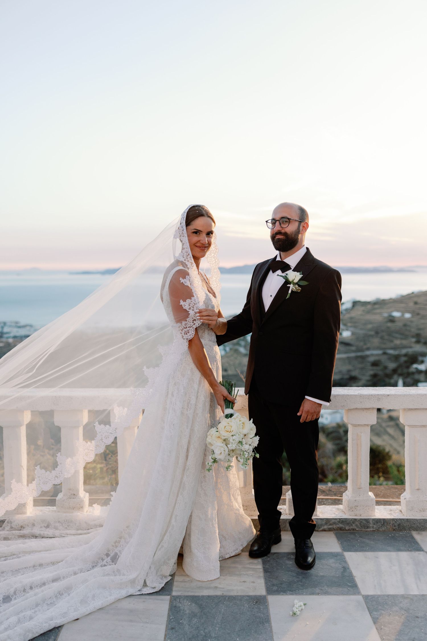Bride in lace gown with flowing veil and groom in tuxedo pose on marble balcony overlooking scenic coastal sunset.