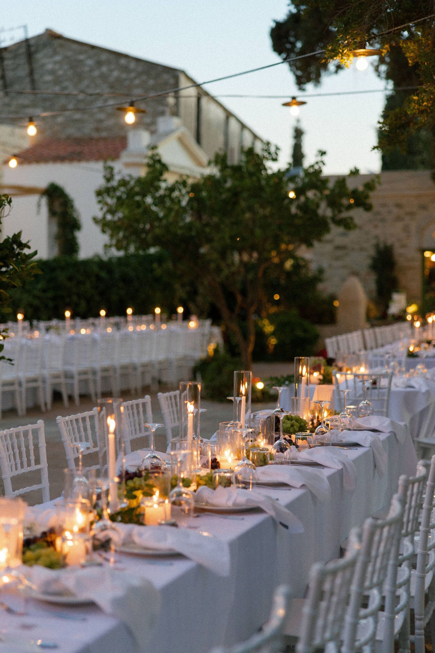 Elegant outdoor wedding reception with long white tables, candles, and string lights at dusk in a Mediterranean courtyard.