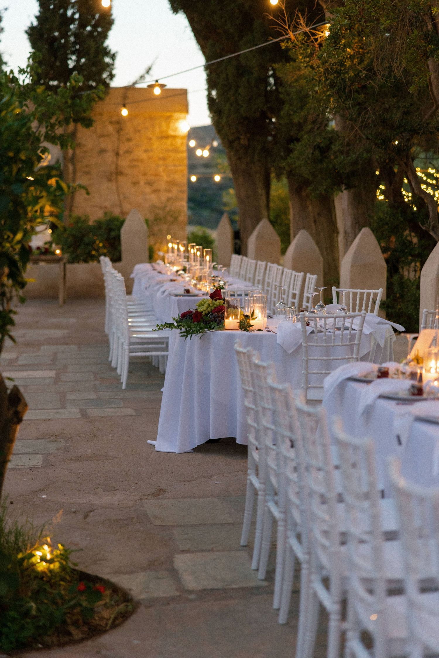 Elegant outdoor wedding reception with long white-draped tables, chiavari chairs, candles, and string lights at dusk.