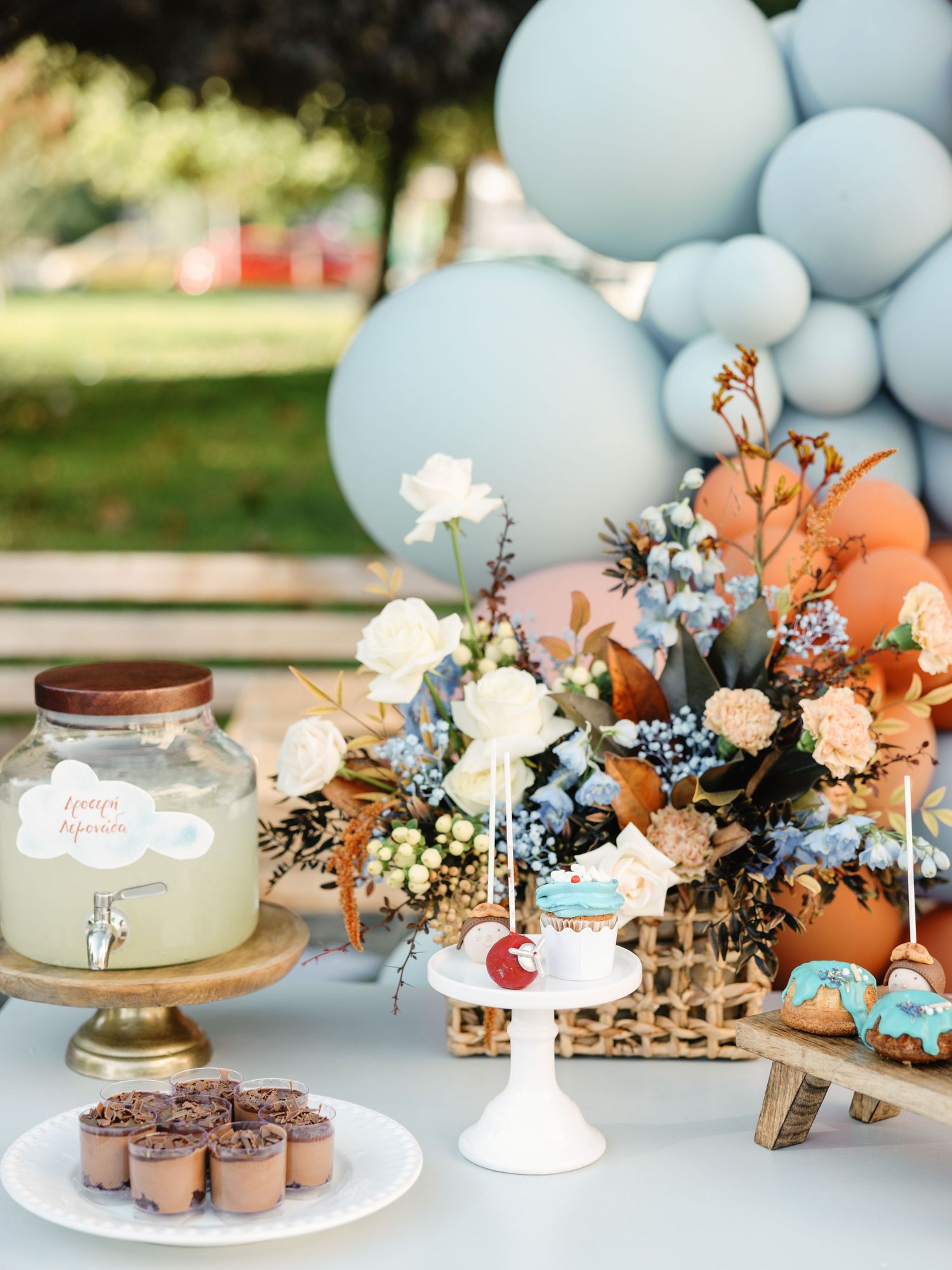 Elegant baby shower dessert table with pale blue balloons, floral arrangements, and chocolate treats on white cake stands.