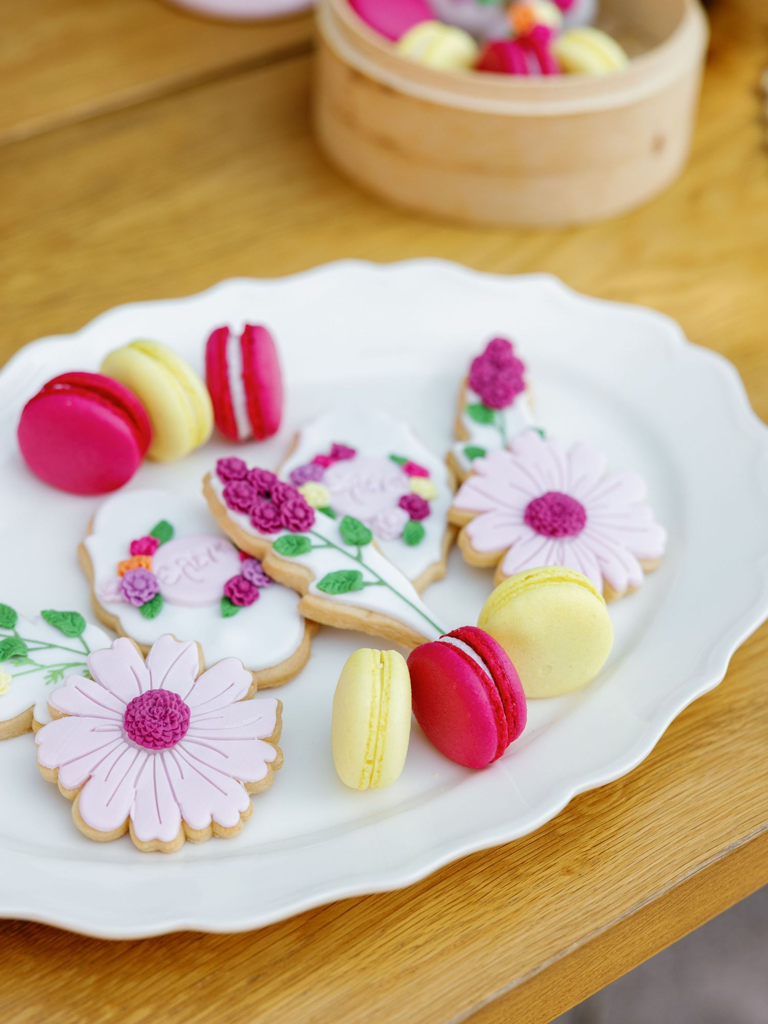 Pink and white flower-shaped sugar cookies with macarons arranged on white scalloped plates against a wooden surface.