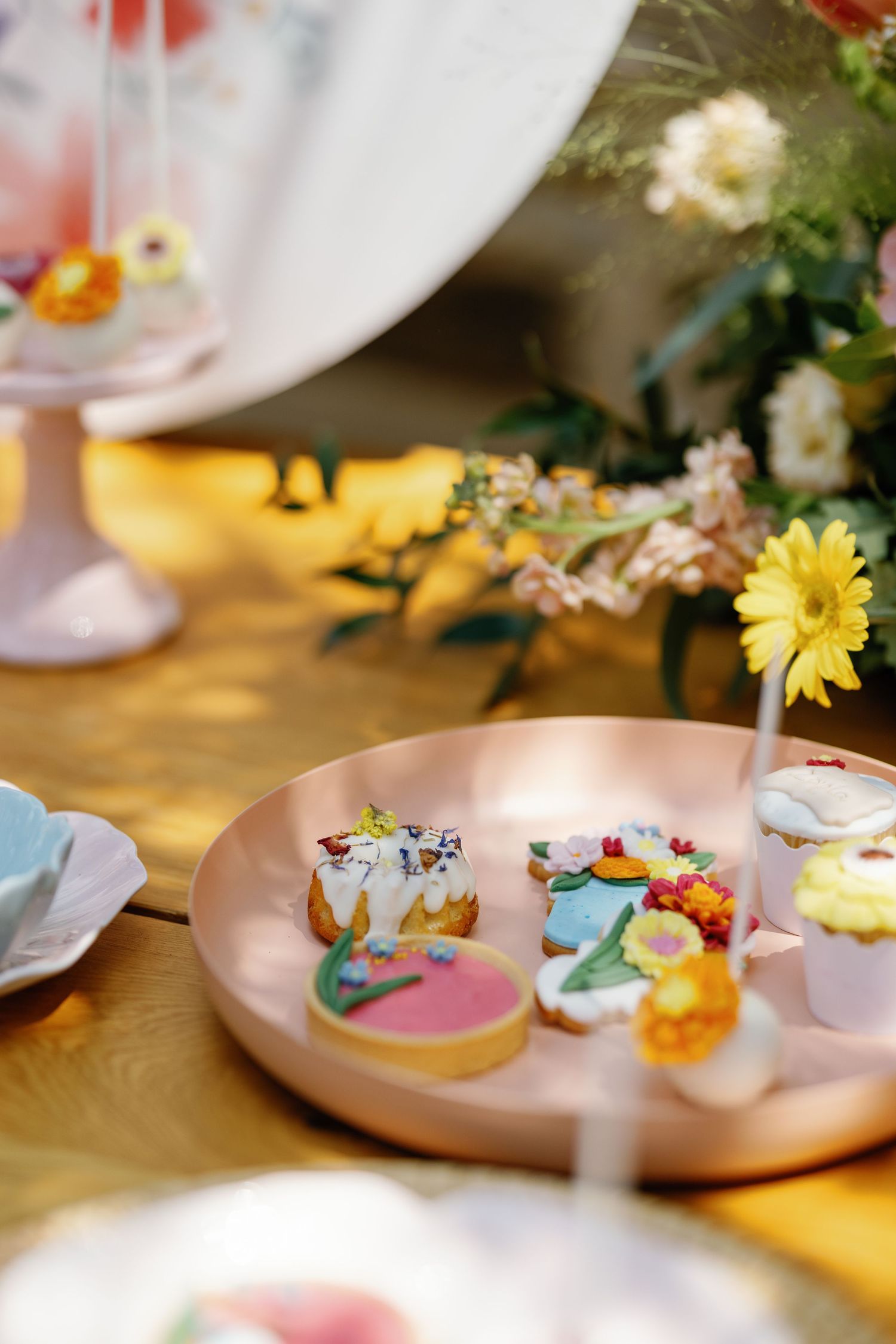 Pink ceramic plates with decorated cookies displayed among fresh flowers on wooden table.