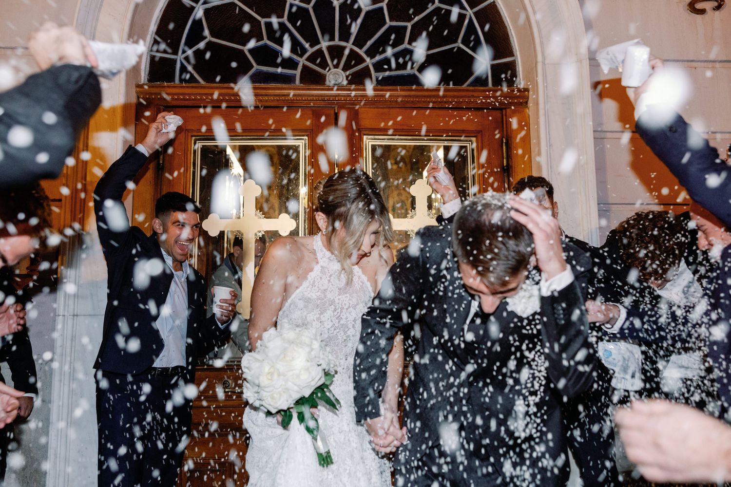 Wedding celebration with guests throwing white confetti under ornate architectural details.