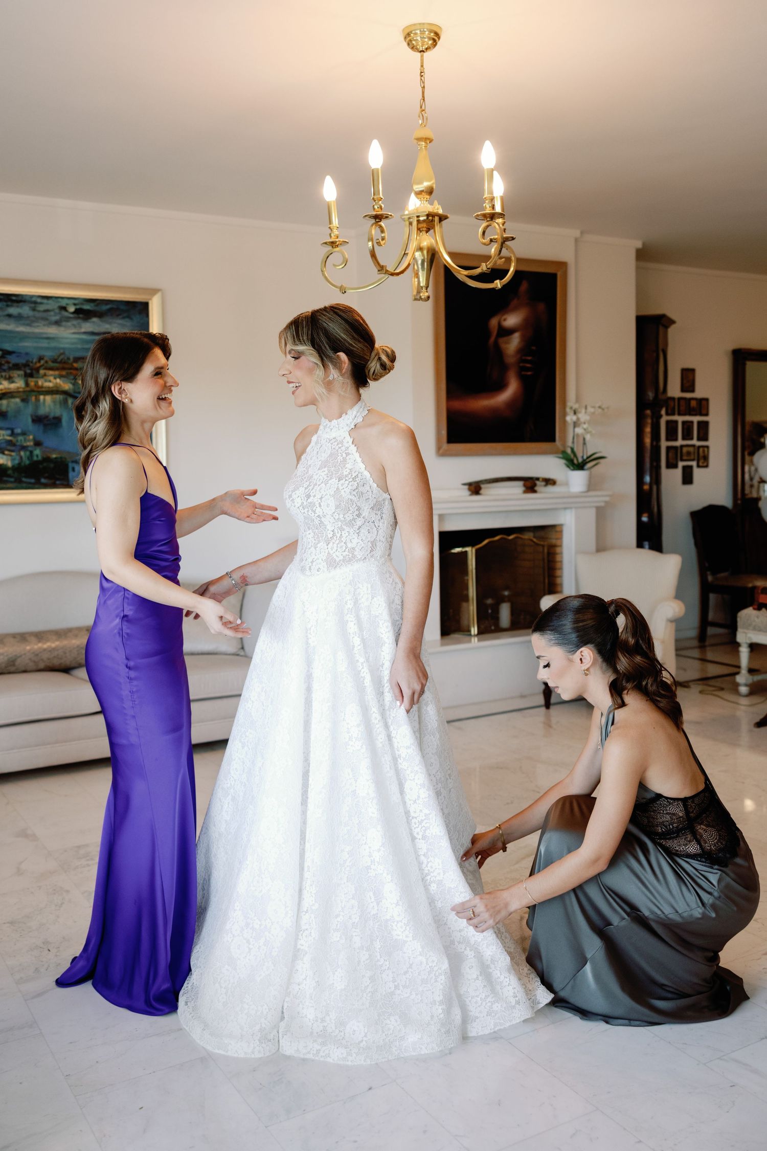 Bridesmaids in purple and gray dresses helping adjust a white wedding gown.