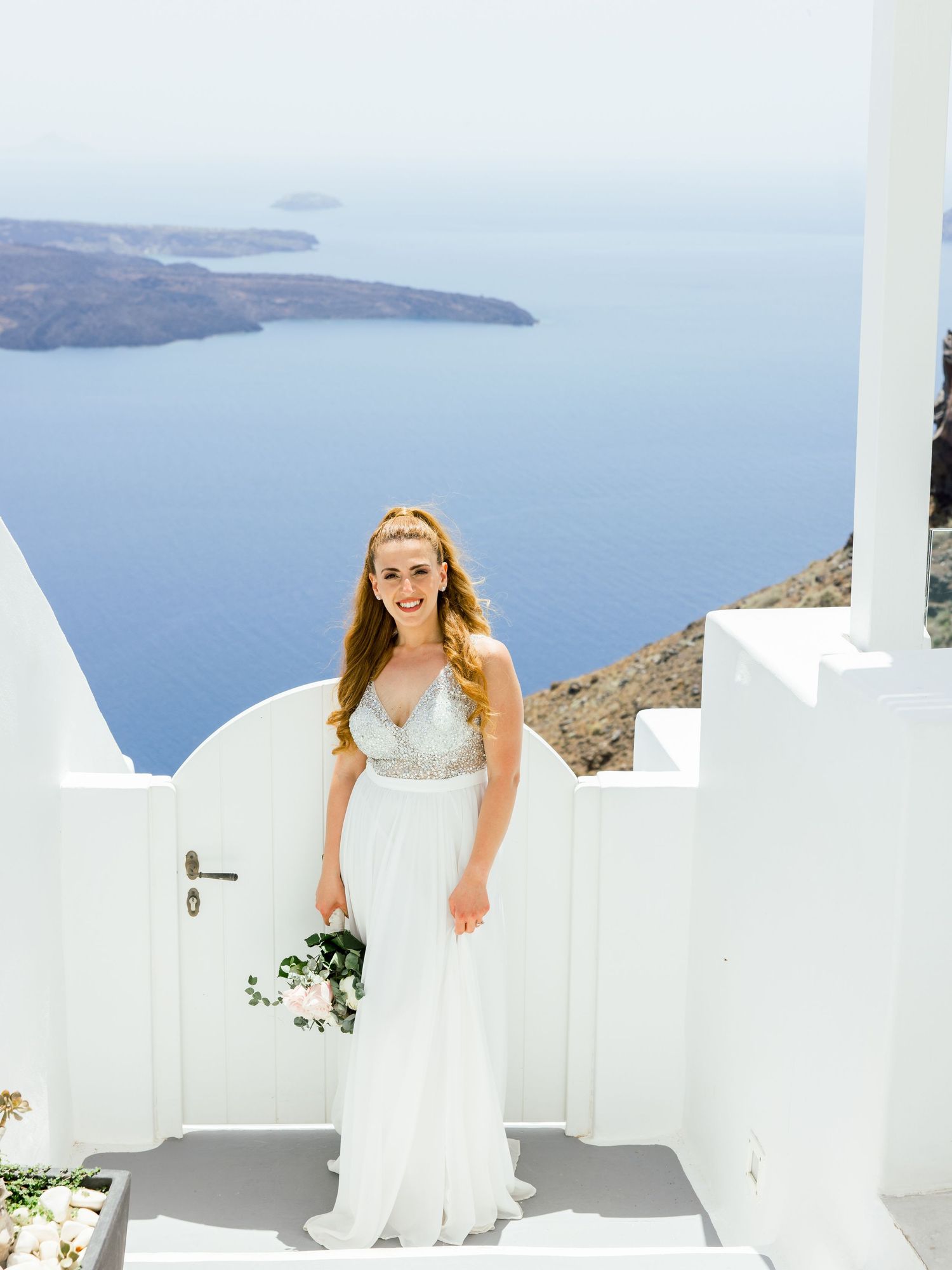 A bride in a flowing white dress stands on white steps with ocean views in Santorini.