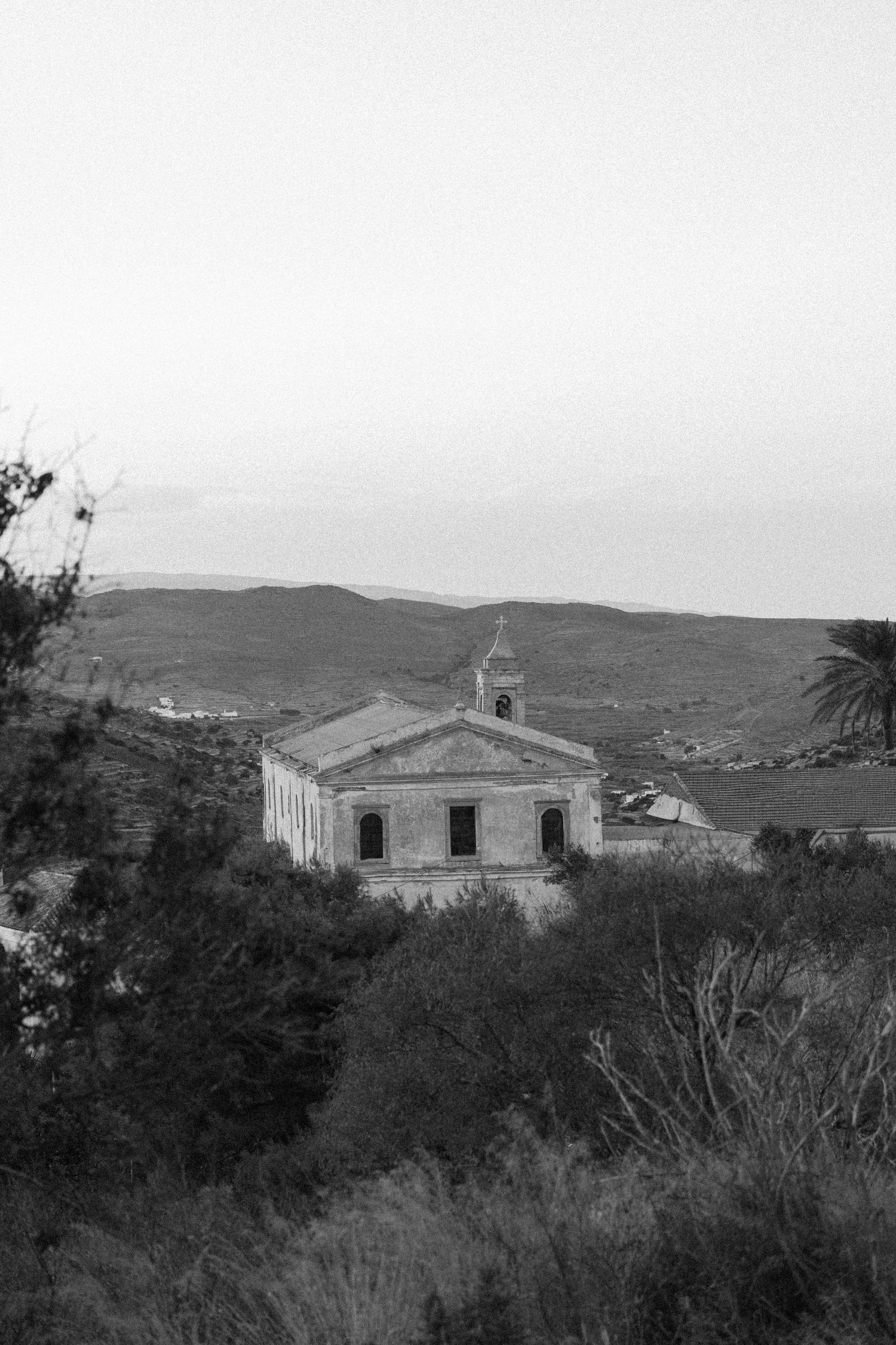 Black and white photo of a historic stone church with bell tower nestled among hills and trees near the sea.