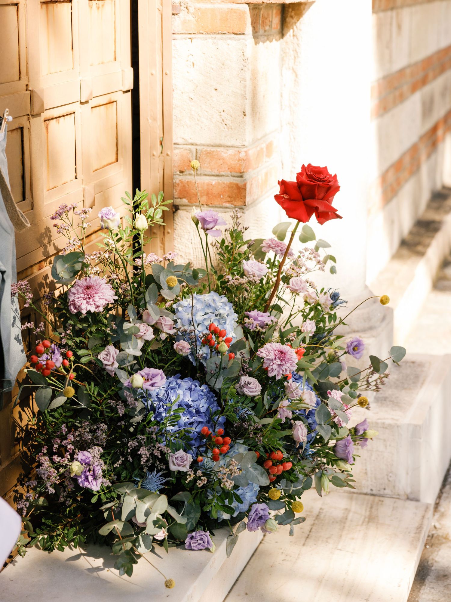 A vibrant floral arrangement with red roses, blue hydrangeas, and mixed wildflowers sits against a sunlit stone wall.