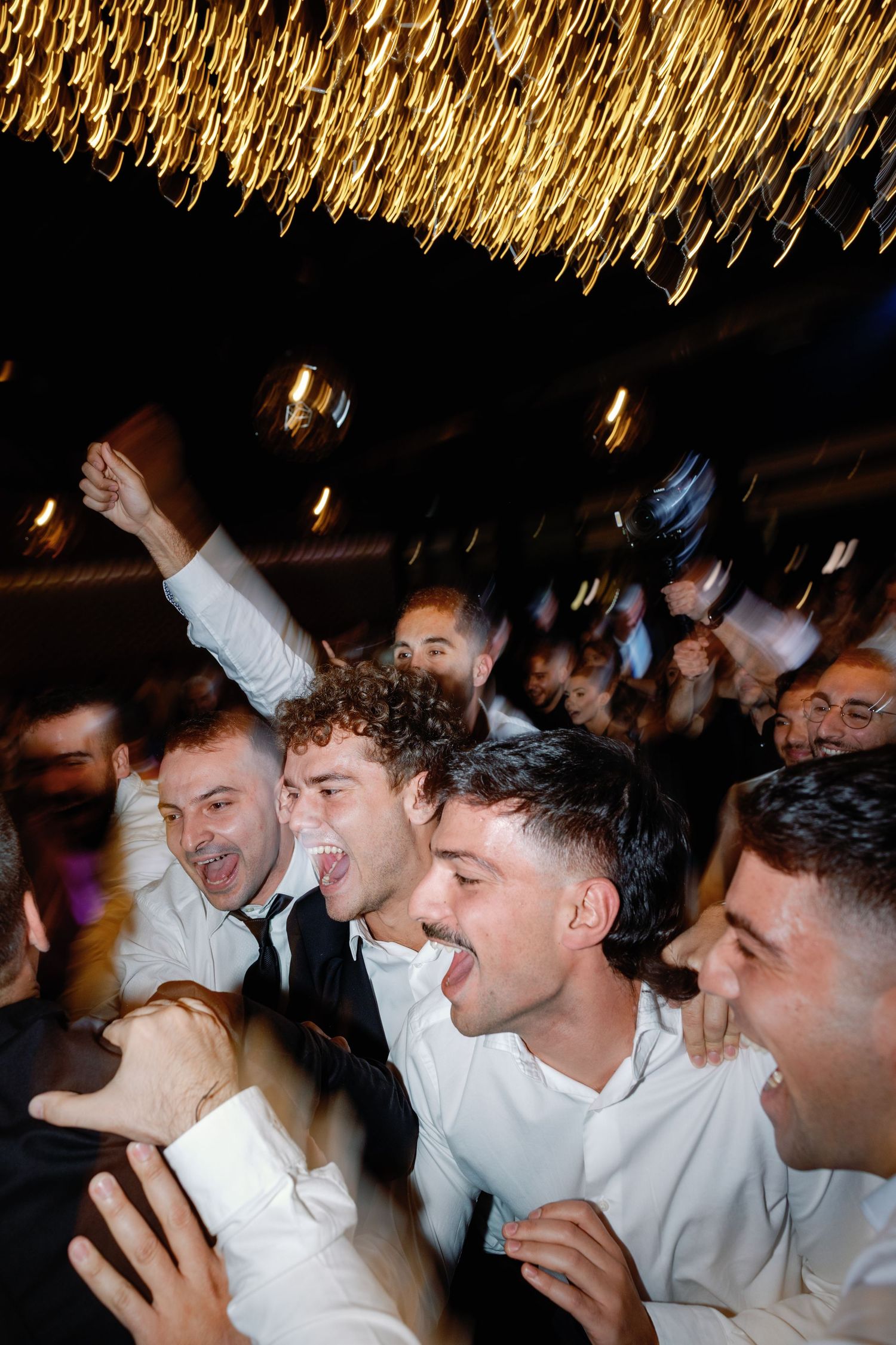 Group of wedding guests in white shirts celebrating under golden string lights.