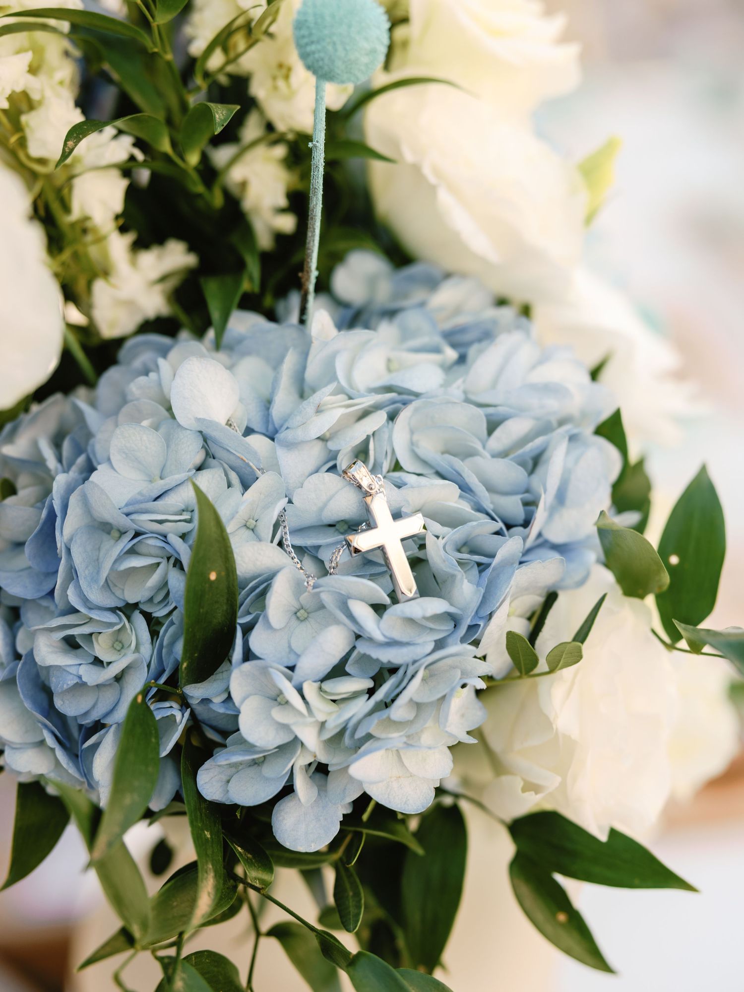 Light blue hydrangeas and white roses arranged with greenery and a small cross decoration for a religious celebration.