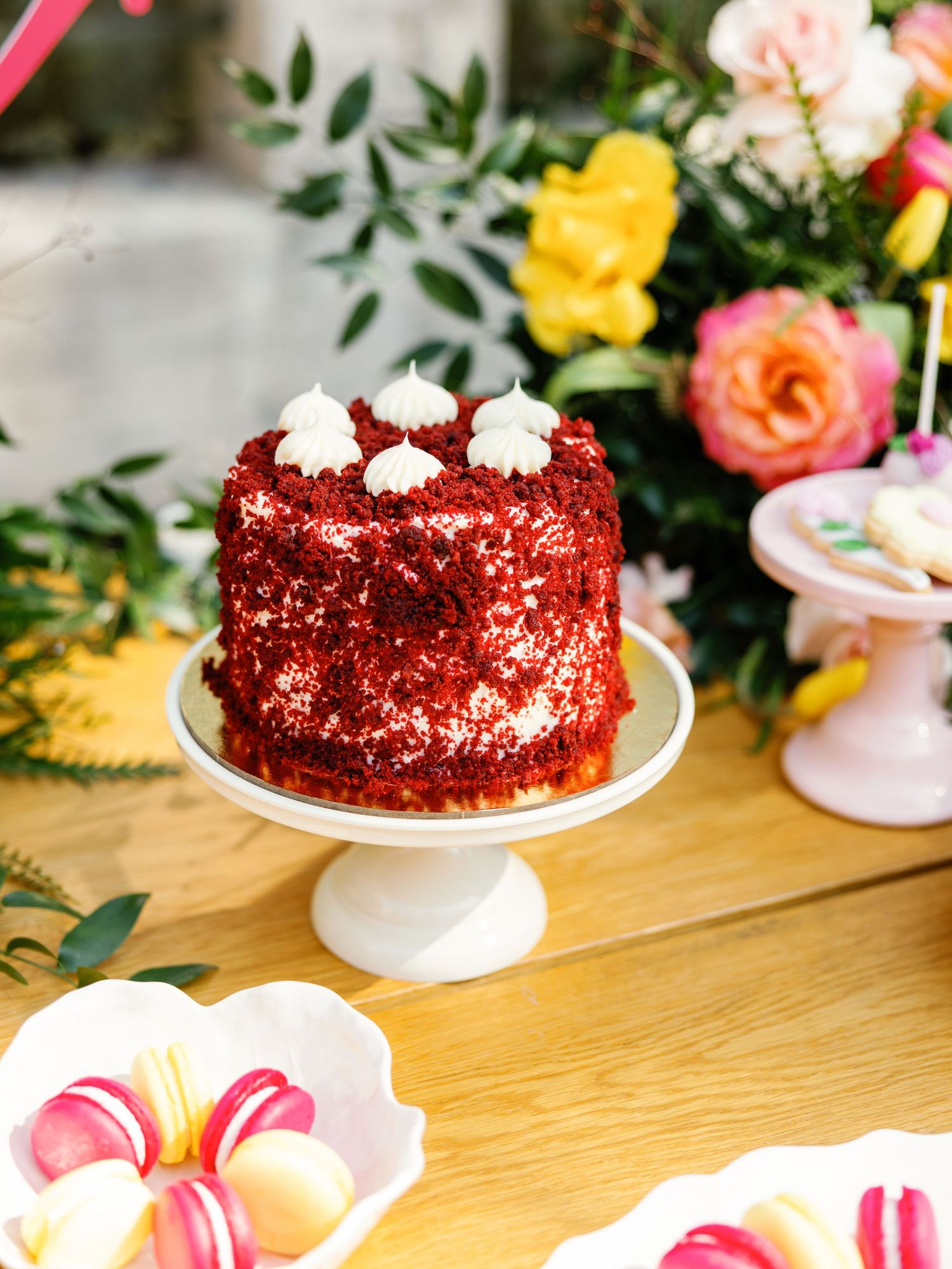 A red velvet cake with white frosting decorations sits on a white cake stand surrounded by colorful roses and flowers.