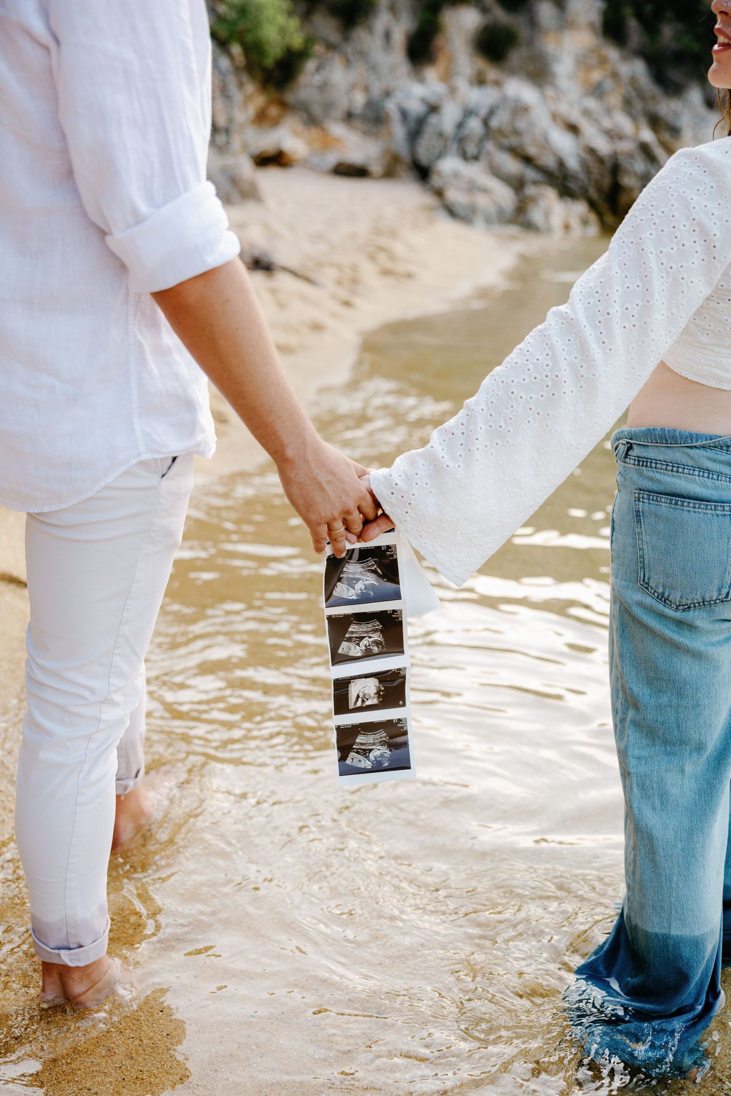 Couple holding ultrasound photos while standing in shallow beach water.