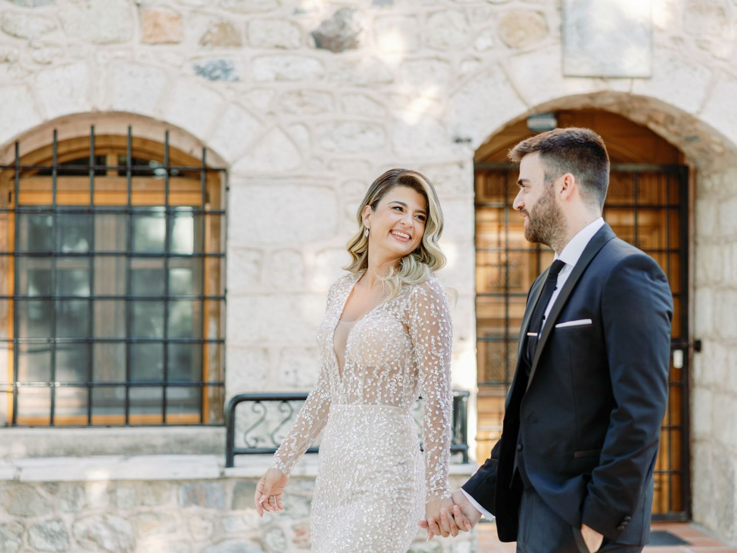 Elegant couple poses against rustic stone wall with arched windows during wedding photoshoot.