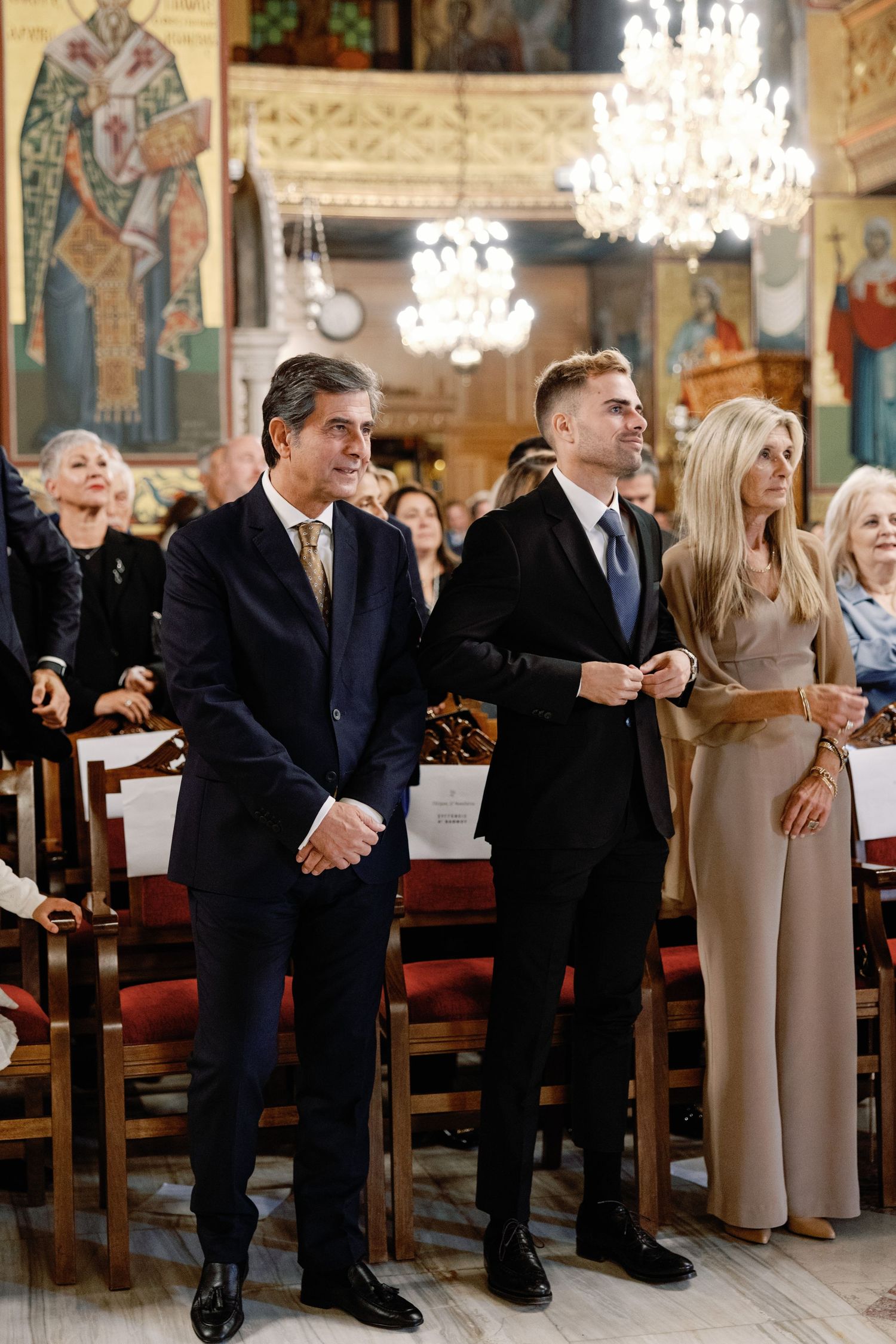 Wedding guests seated in elegant church pews during traditional ceremony with ornate chandeliers overhead.