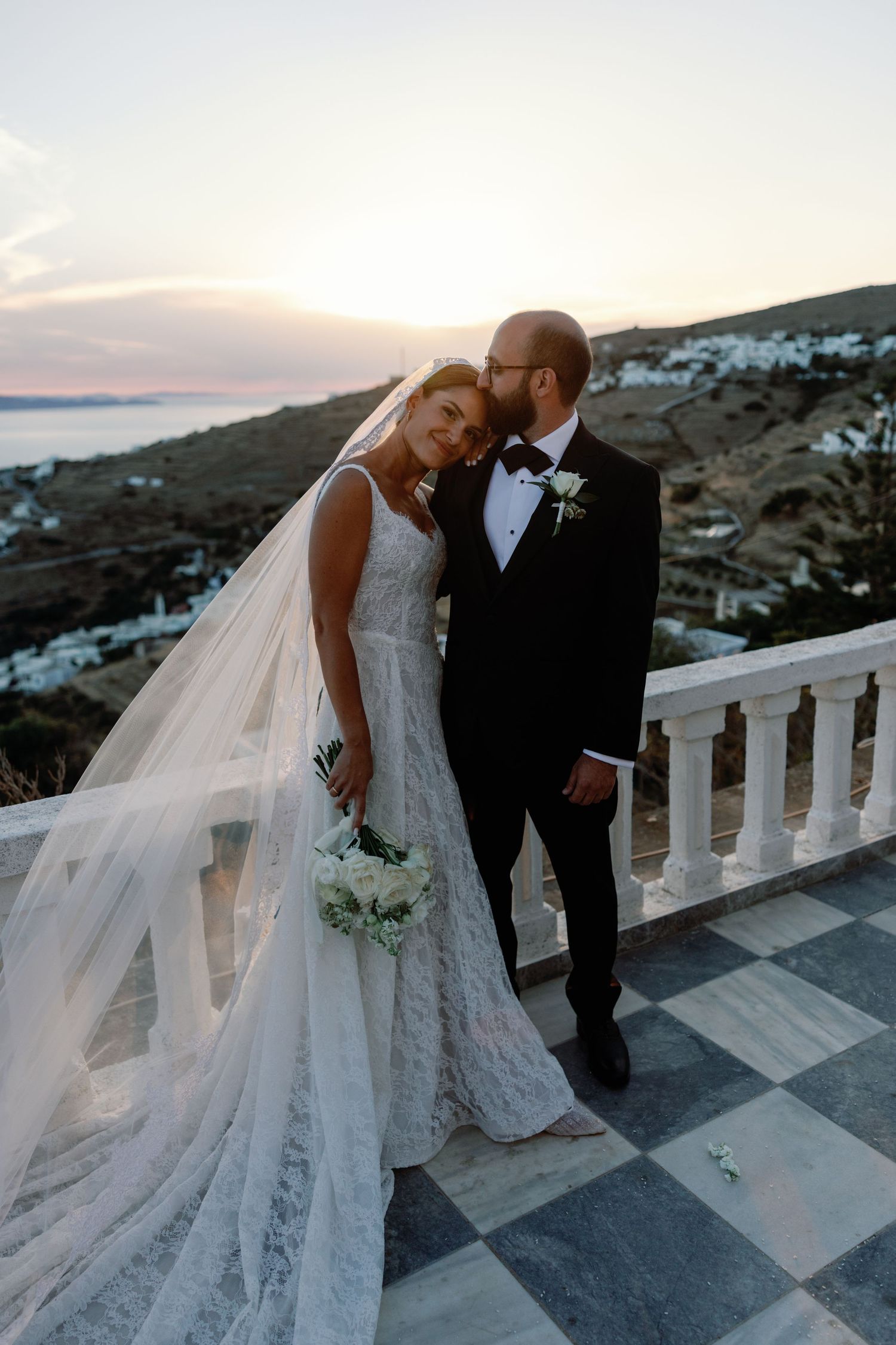 Bride and groom share a romantic sunset moment on a terrace in Santorini, Greece, with white buildings and sea views.