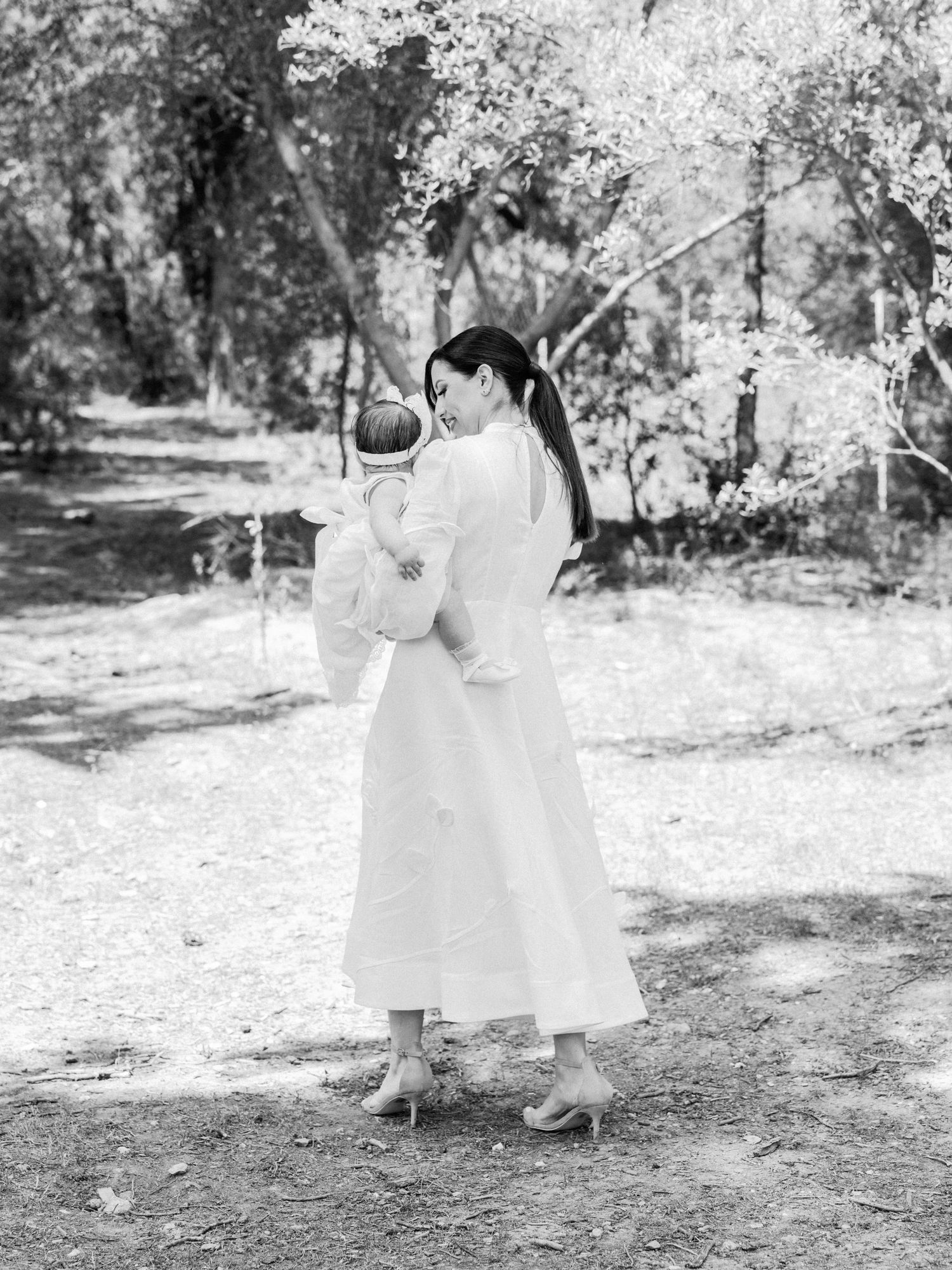 Two young children in white dresses walk together through a sunlit wooded path in a candid black and white photo.