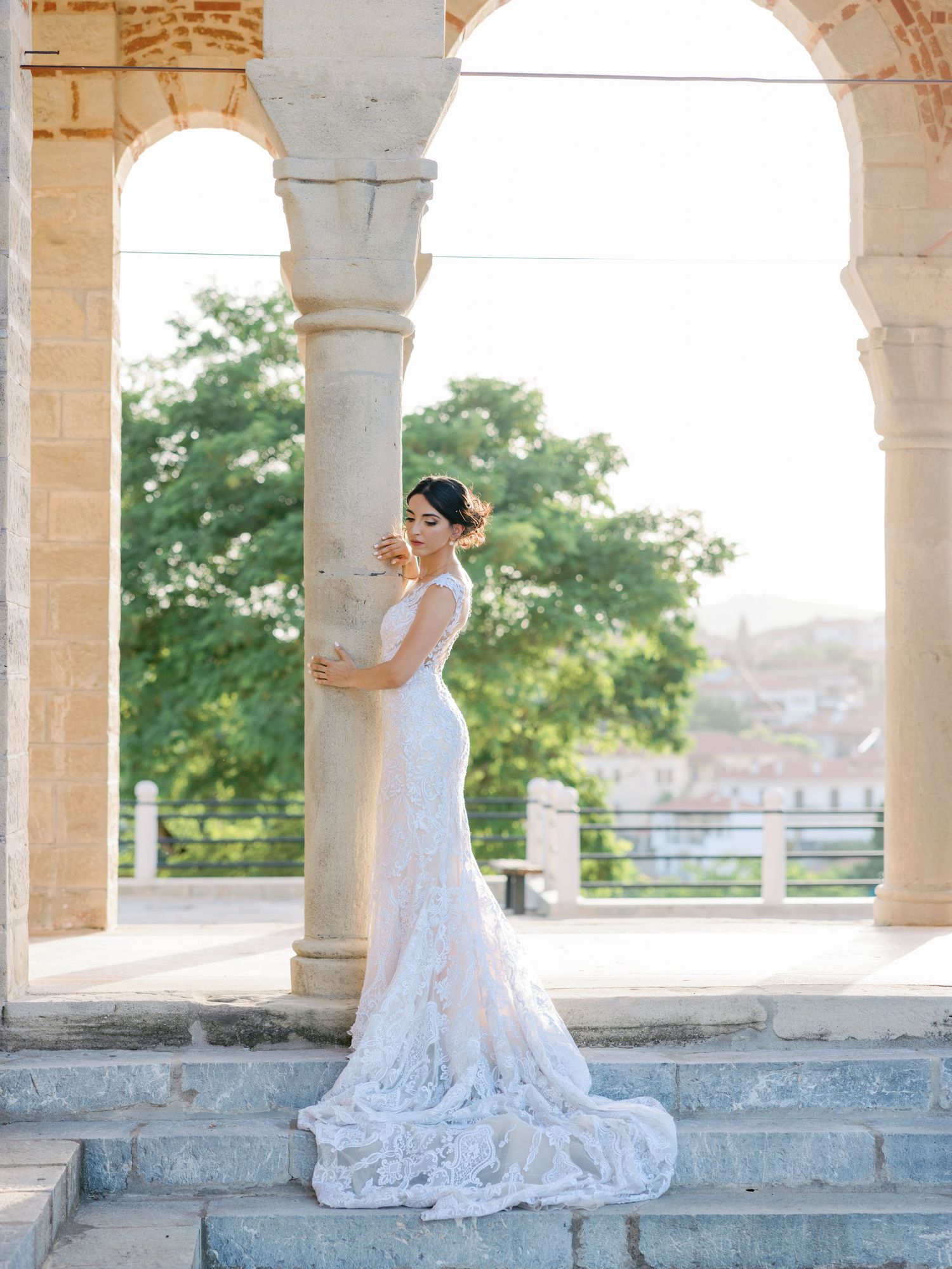 A bride in a lace gown with long train poses elegantly under stone archways.