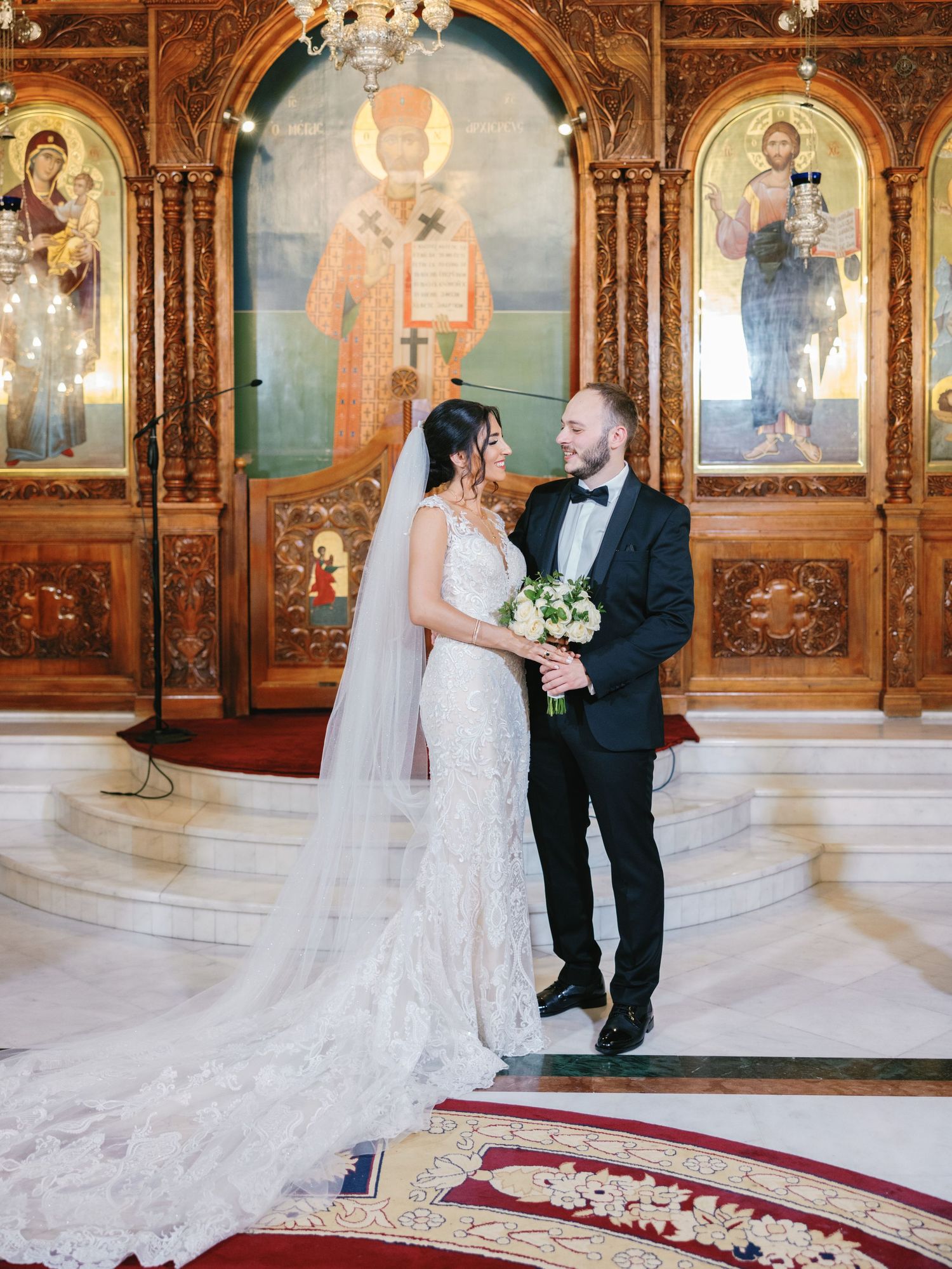 A wedding couple stands before an ornate Orthodox iconostasis with beautiful religious artwork and red carpet.