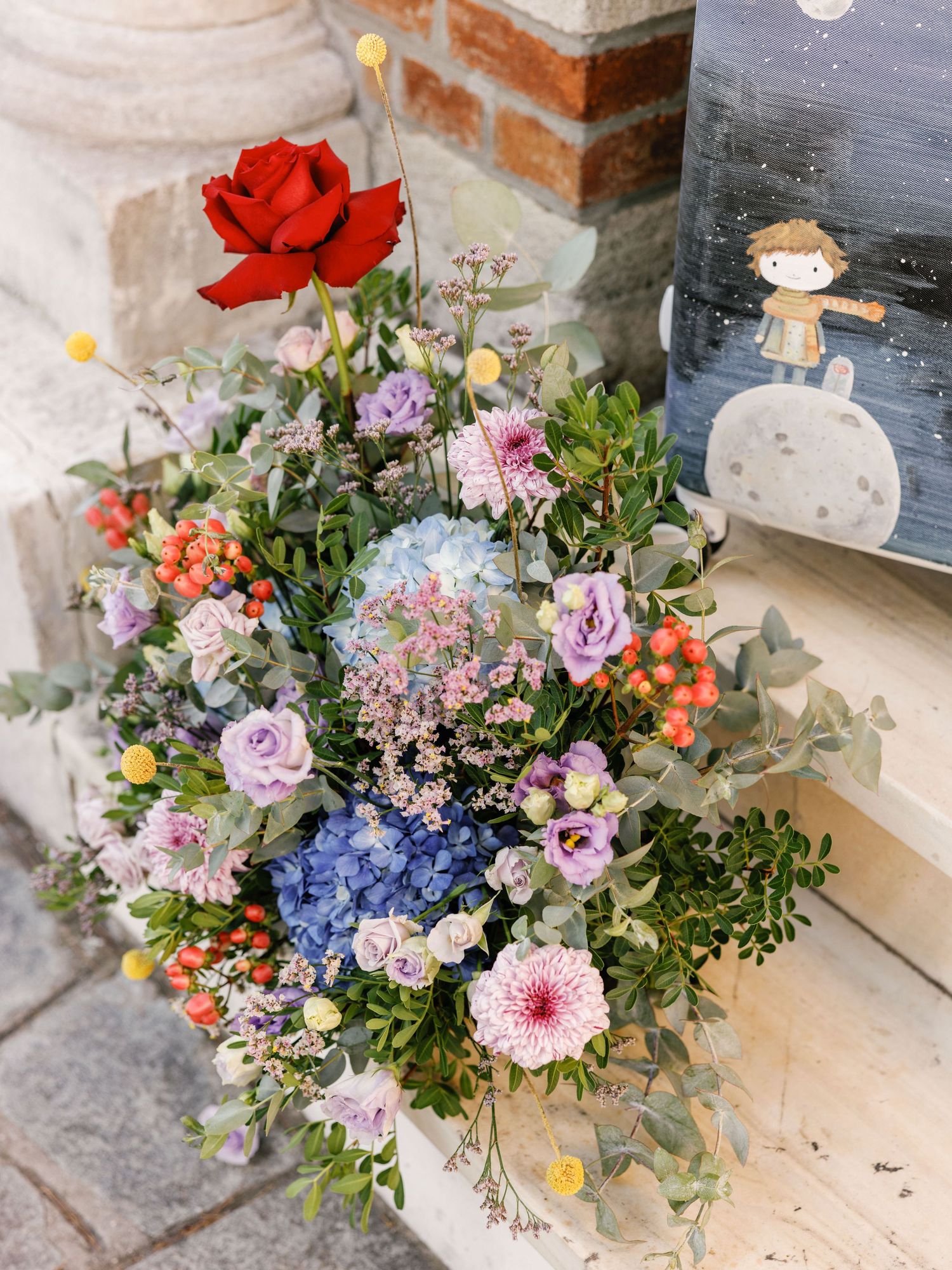 A vibrant floral arrangement with roses, hydrangeas, and mixed blooms in pink, blue, and coral tones beside a framed artwork.