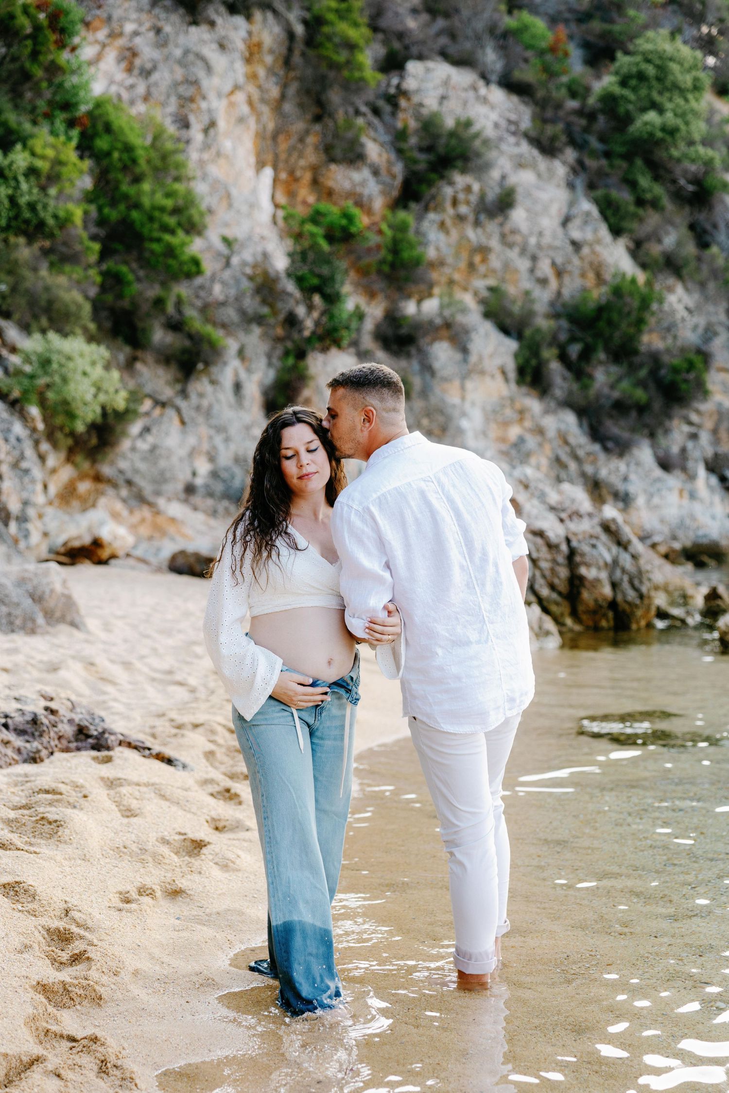 Couple shares intimate moments during beach maternity photoshoot near rocky cliffs.