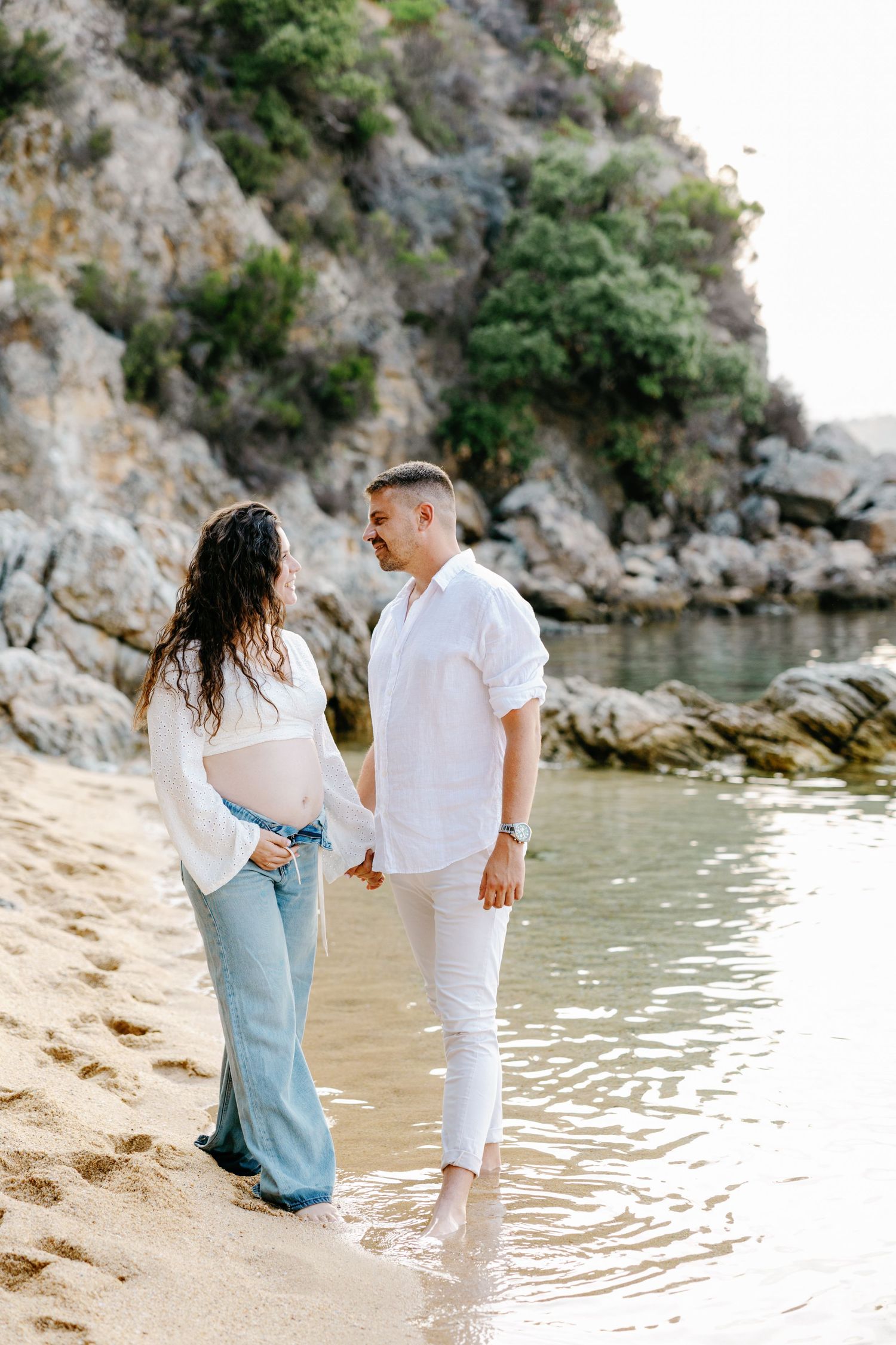 Romantic couple walking along shoreline in matching white outfits during beach photoshoot.