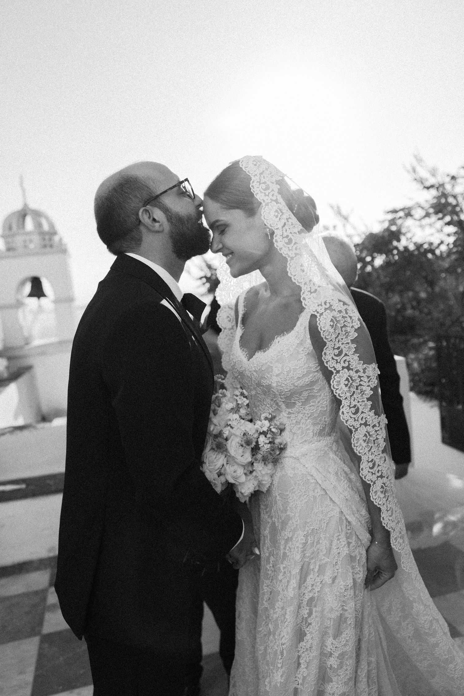 Black and white wedding photo of bride in lace veil and groom in tuxedo sharing an intimate moment outdoors.