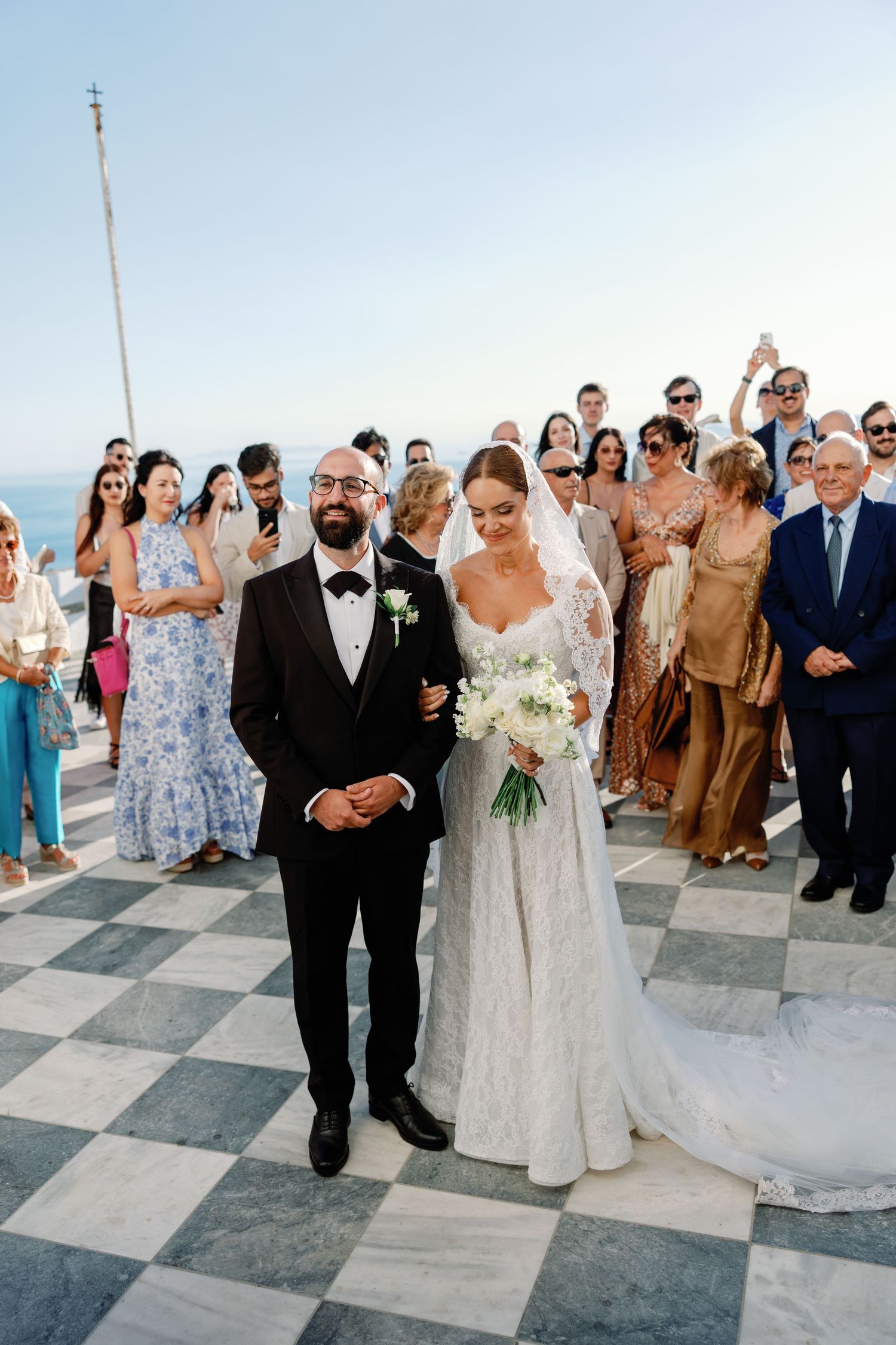 Bride and groom walk down checkered aisle at outdoor wedding ceremony with guests and ocean view in background.