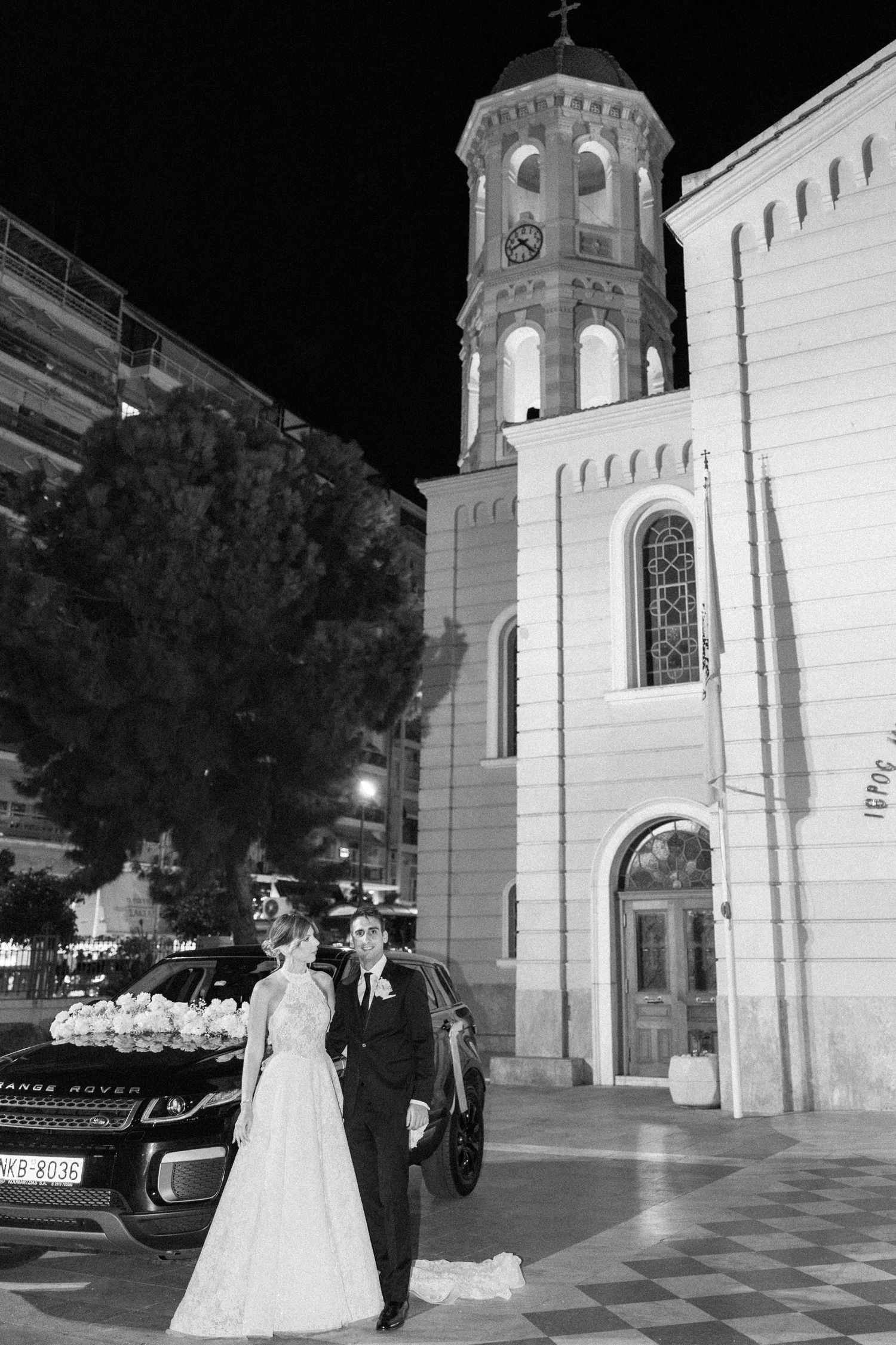 A wedding couple poses outside a grand church at night with illuminated architecture.